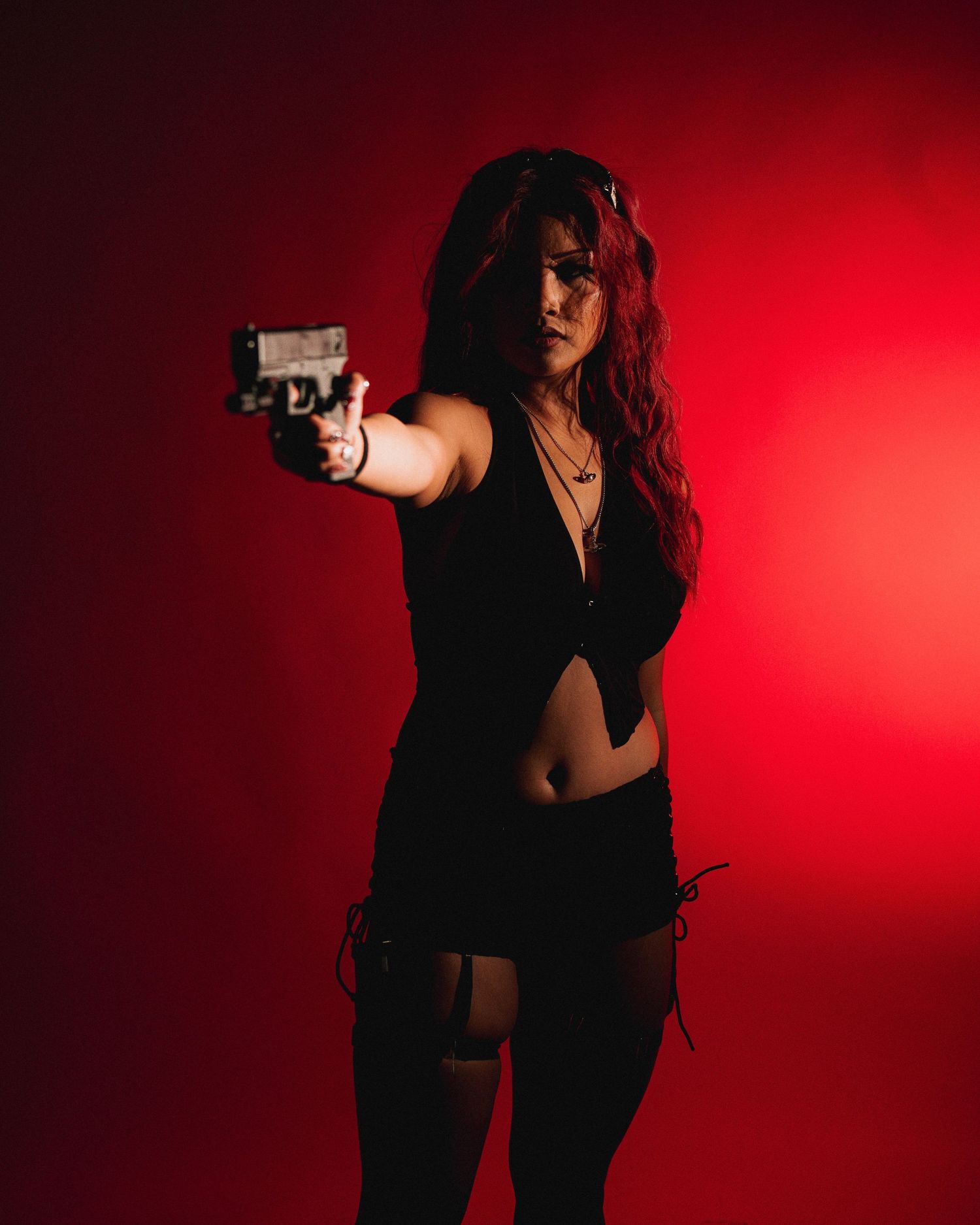A dark-haired figure in black clothing aims a handgun against a dramatic red background.