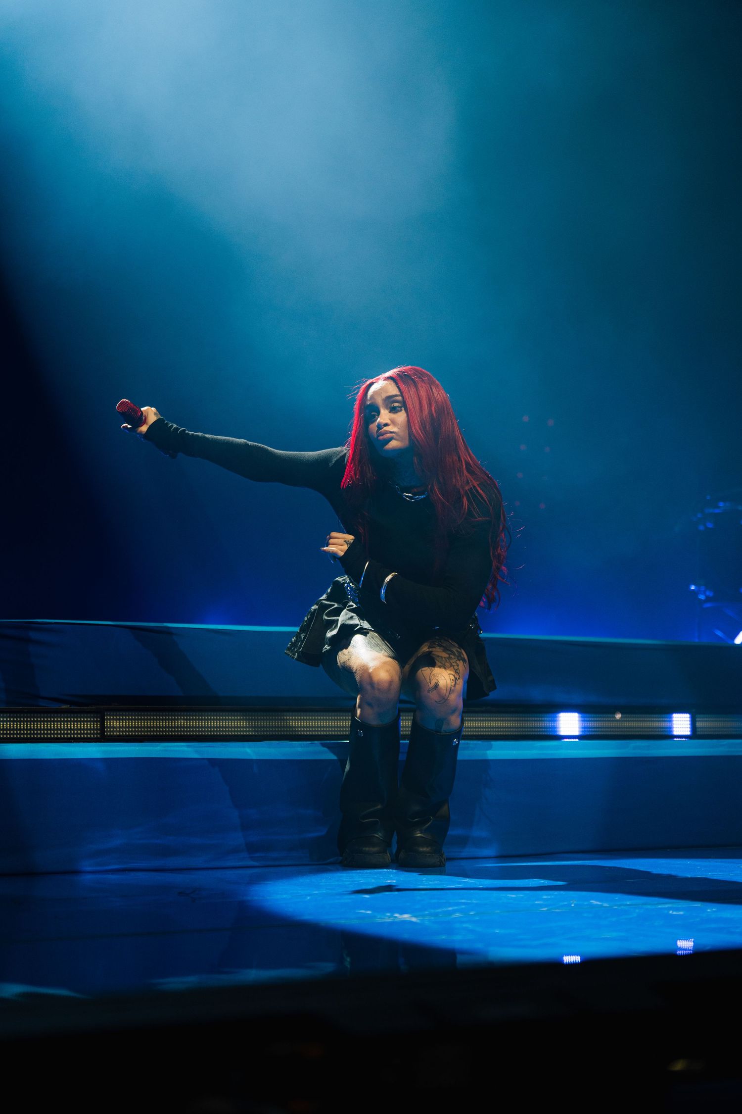 Performer with long red hair in black outfit strikes dramatic pose on darkened stage with blue lighting effects.