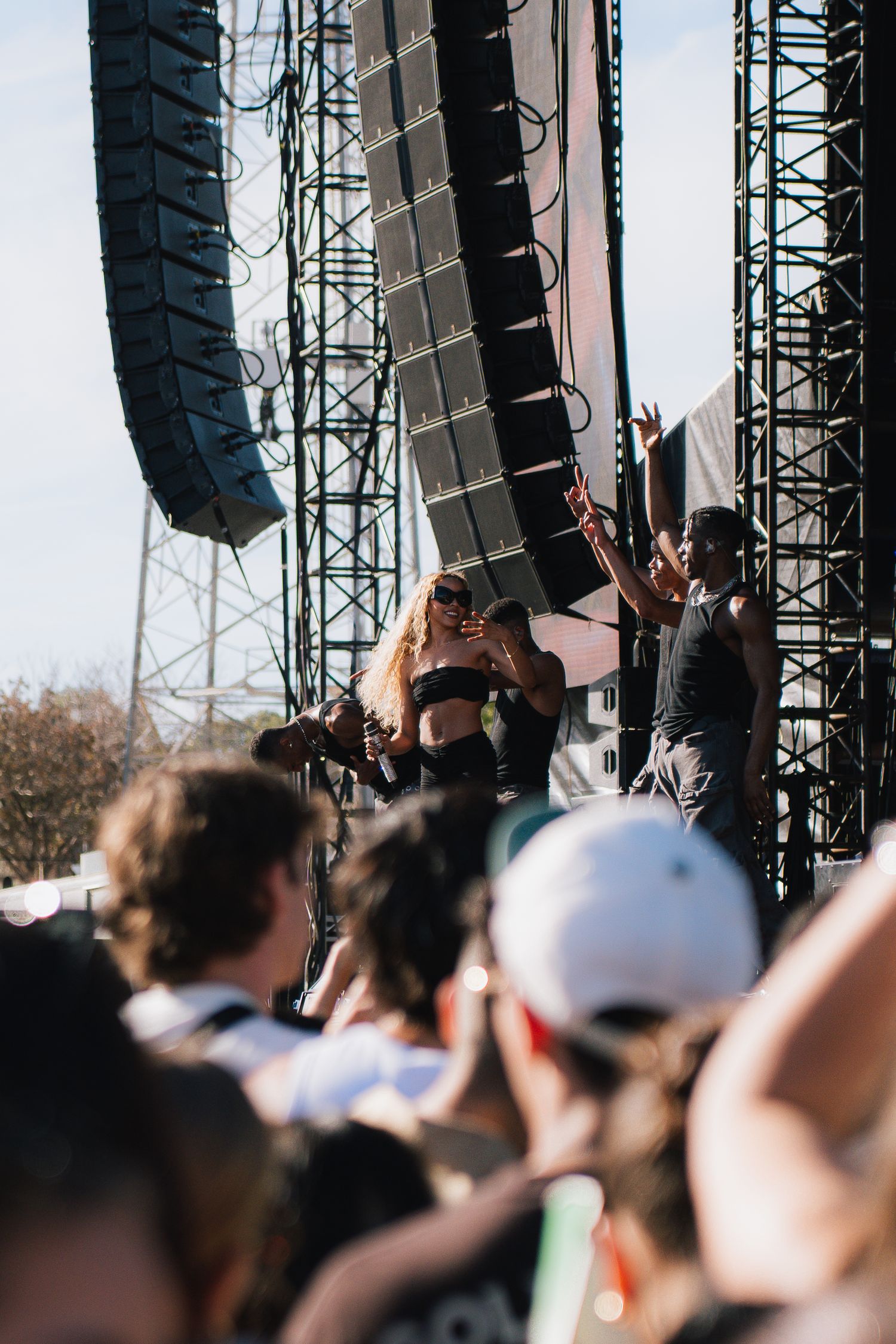 Concert crowd watching stage with large line array speaker system suspended above during an outdoor music festival performance.