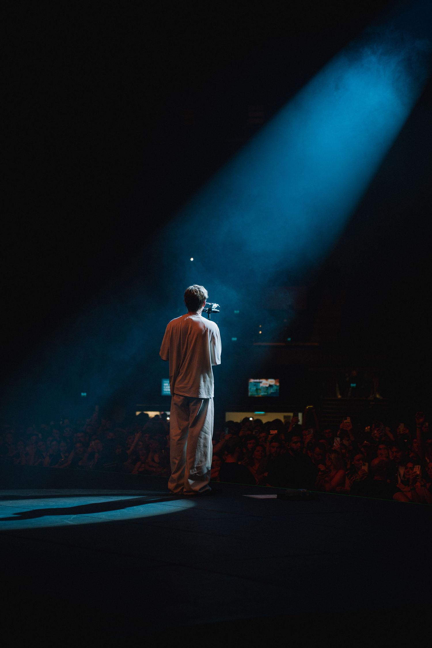 Performer standing alone on stage under dramatic blue spotlight beam while crowd watches from below in dark venue.
