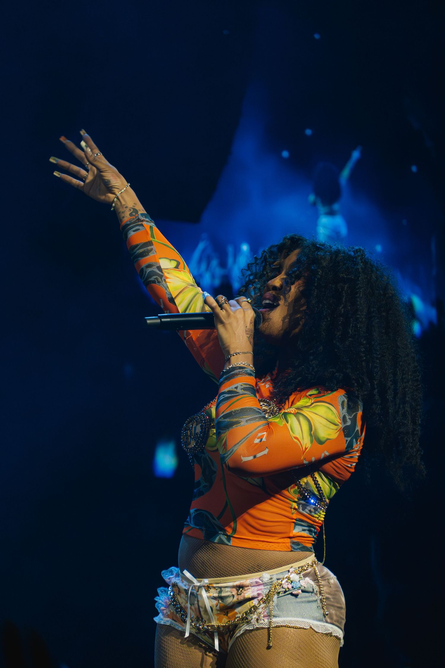 Musical performer in an orange top sings passionately on stage with arm raised under dramatic blue lighting.