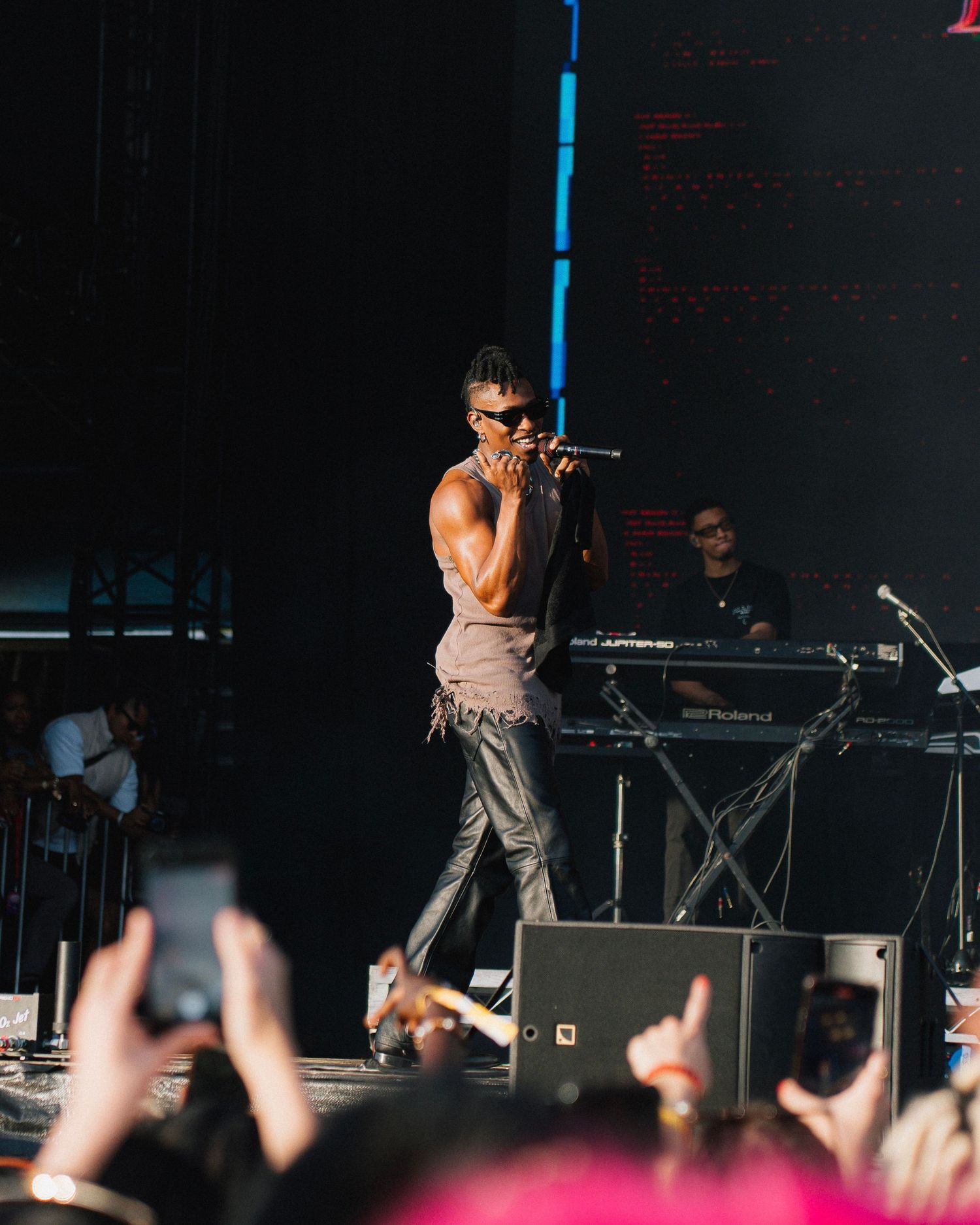 Performer in sunglasses sings on stage at concert while audience members hold up phones to record in dark venue.