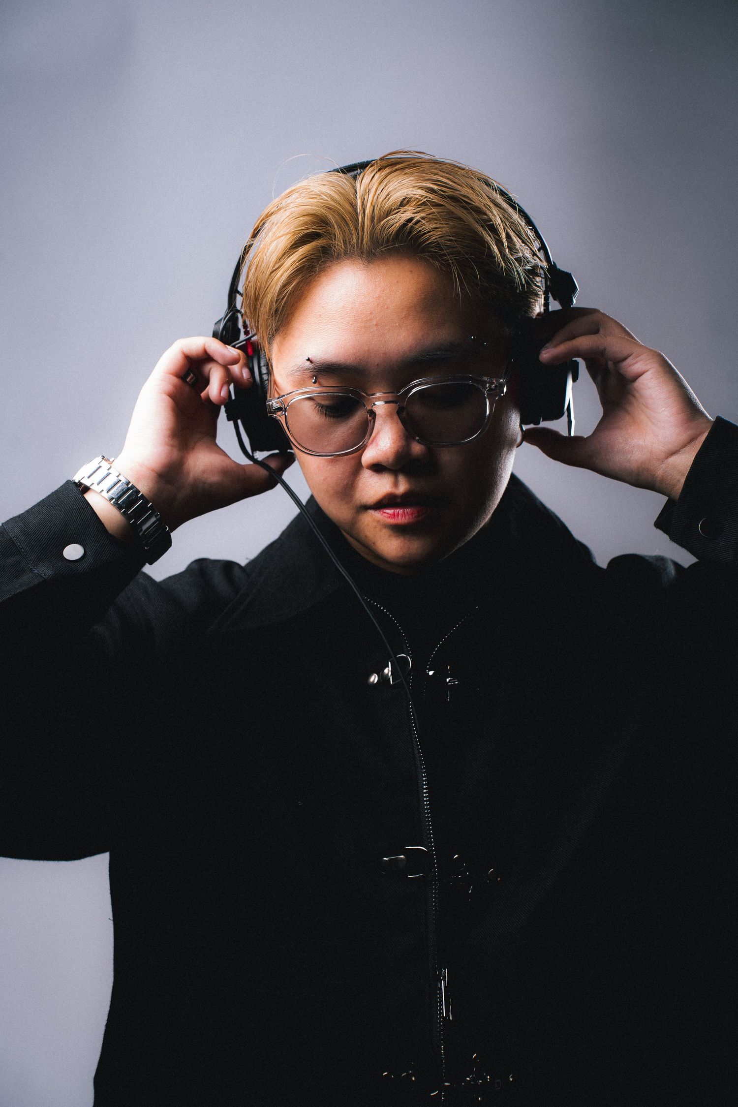 Portrait of someone wearing glasses and headphones in a black jacket with blonde hair against a gray background.