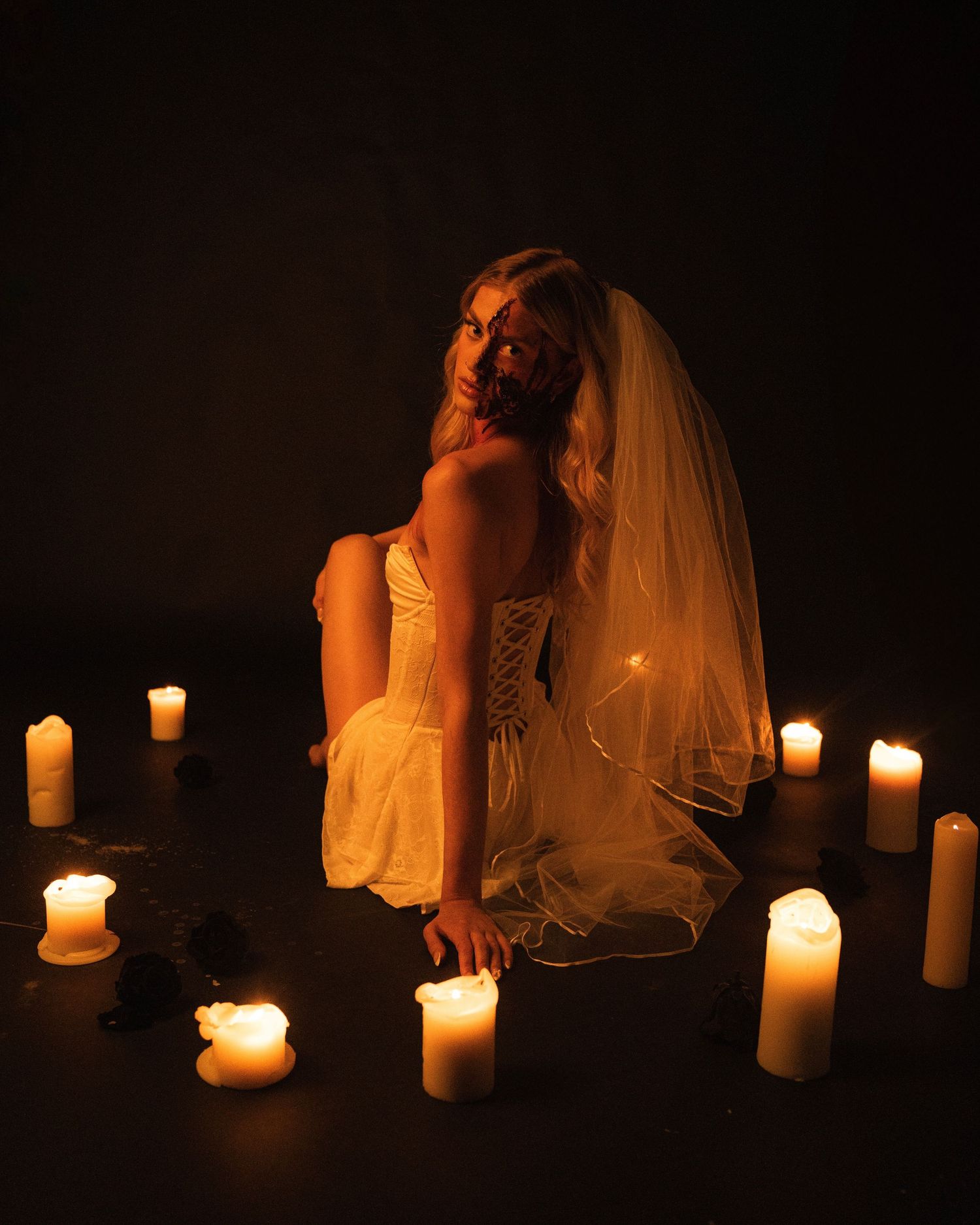 A bride in a white dress and veil kneels dramatically in darkness surrounded by glowing candles.