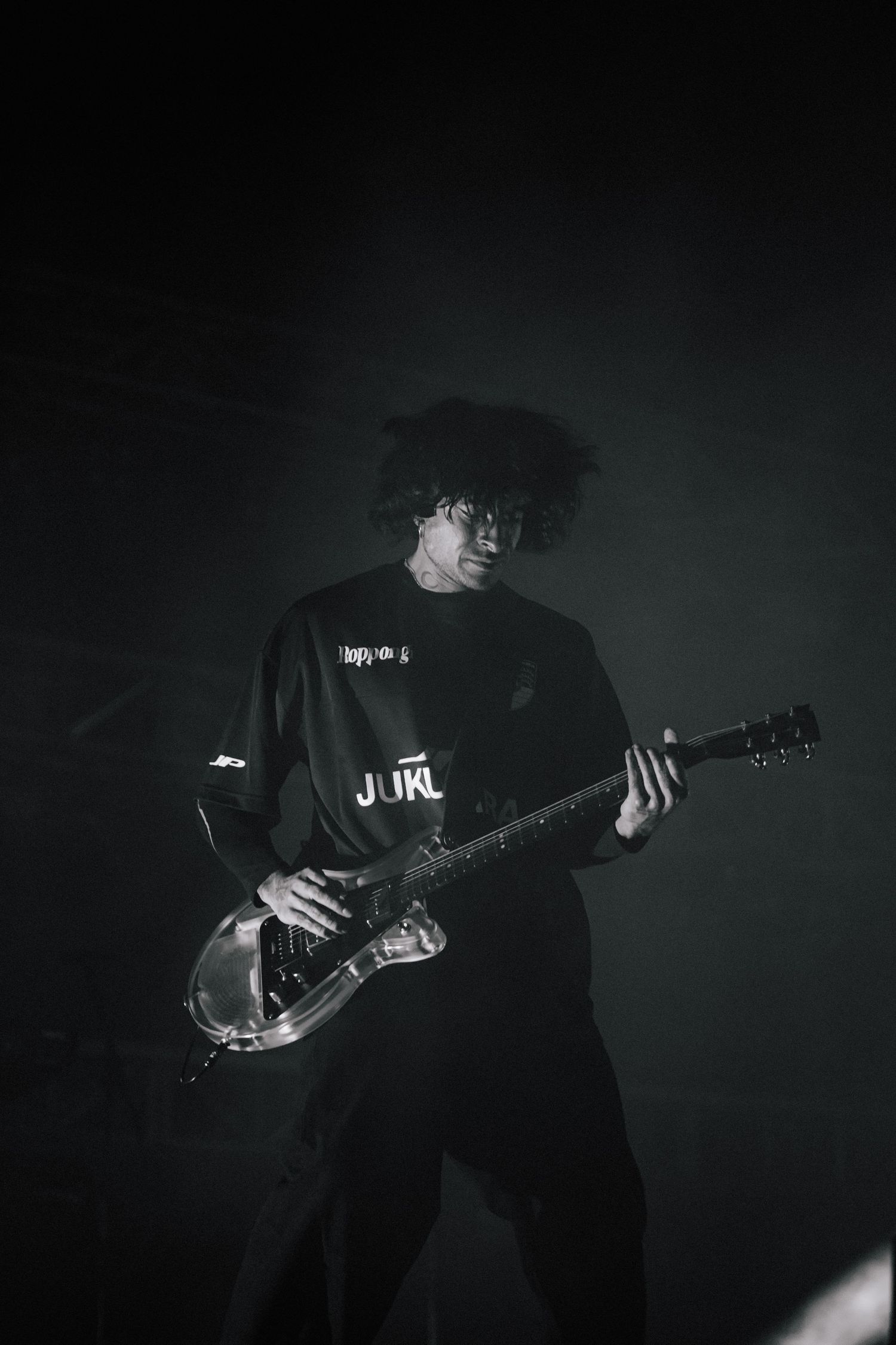 A guitarist in dark clothing plays an electric guitar on stage in moody atmospheric lighting.