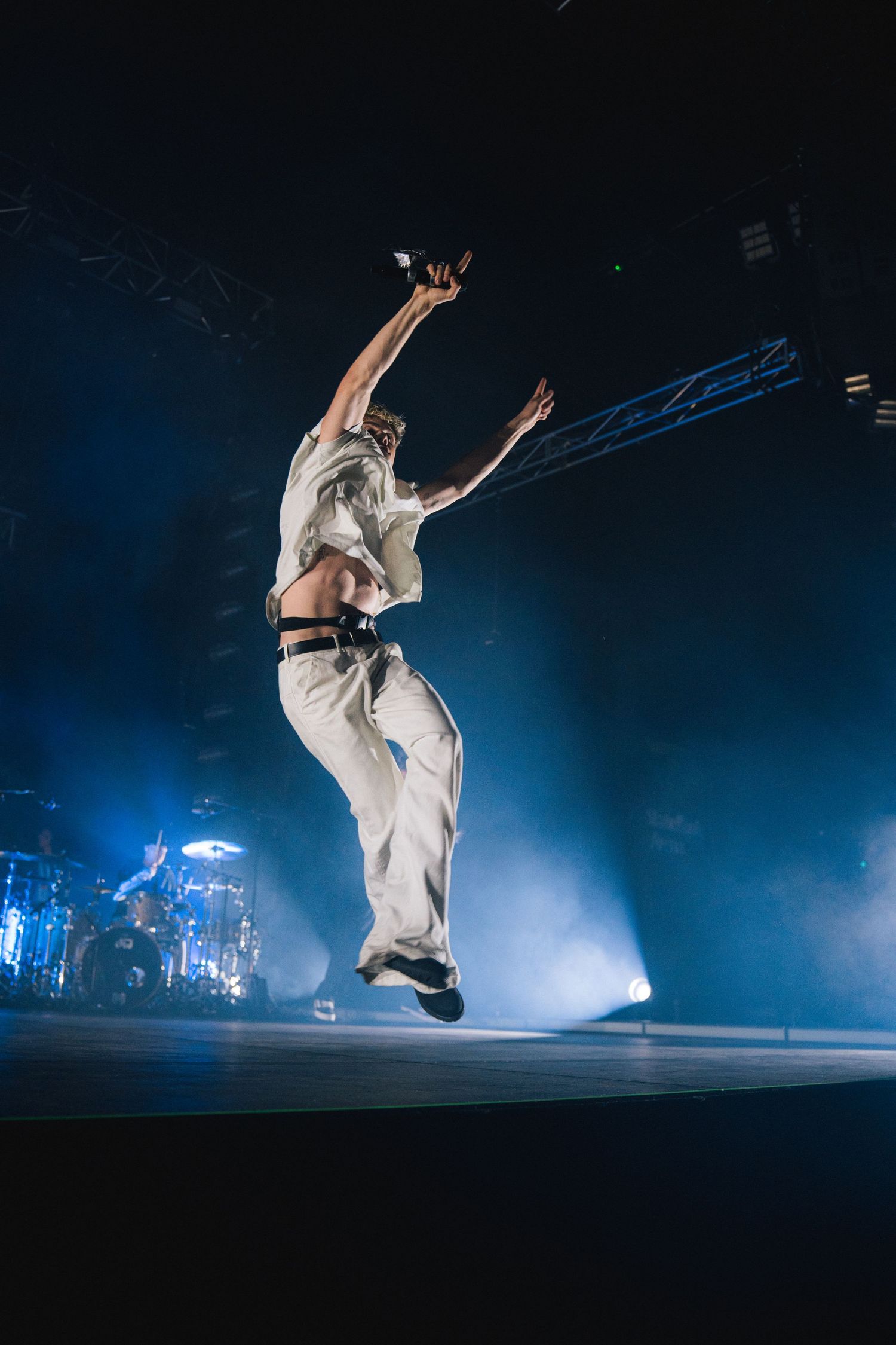 Performer in white outfit leaps dramatically on stage with blue lighting and fog effects during live performance.