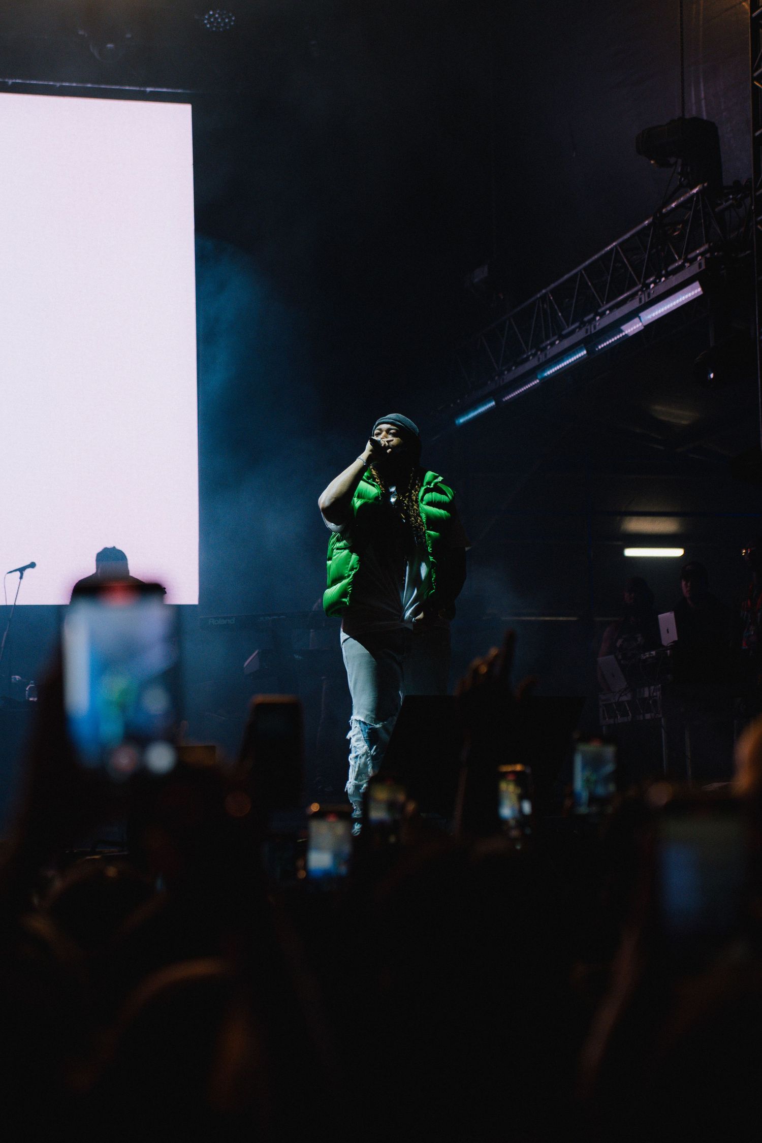 Performer in green illumination jumps on dark stage during concert with crowd and phone lights visible below.