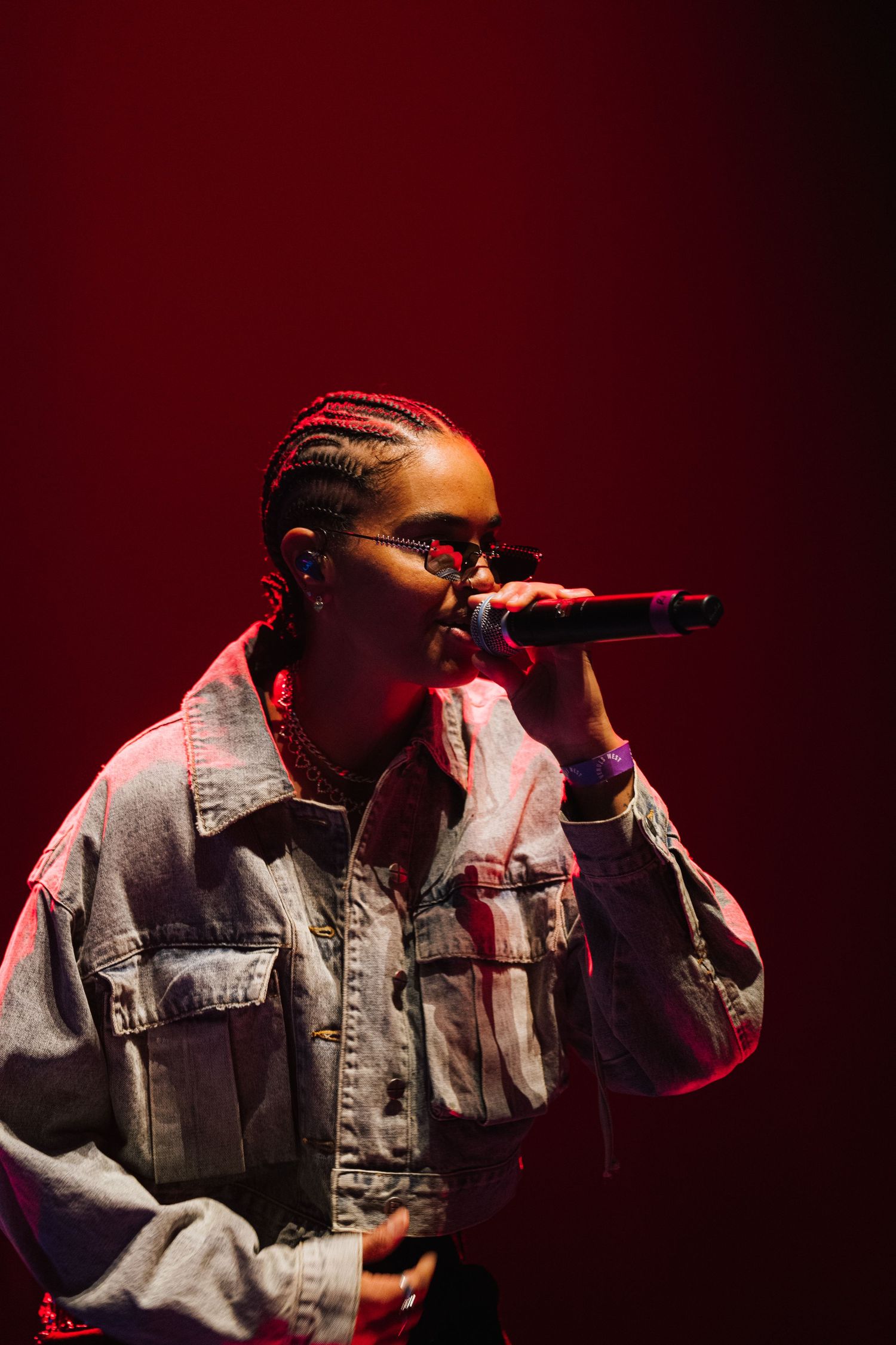 A performer in a denim jacket holds a microphone while singing on stage with dramatic red lighting in the background.