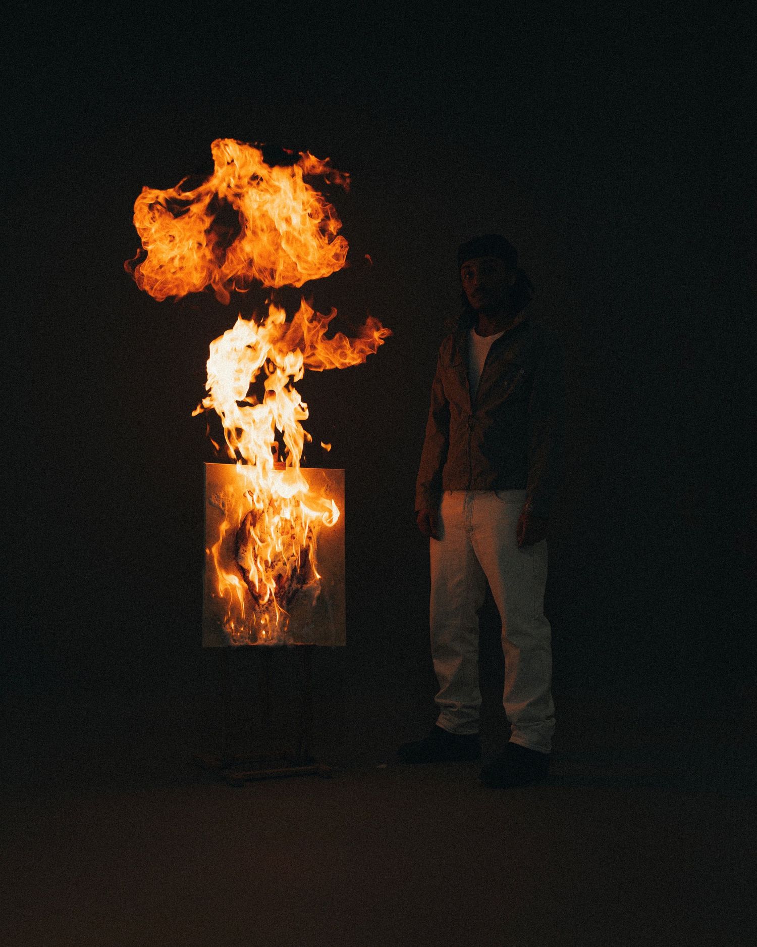 Figure standing next to dramatic fire display in dark room with burning barrel creating intense flames.