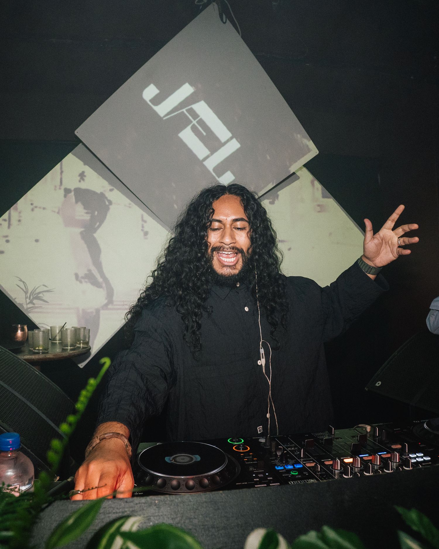 DJ performing energetically behind decks with geometric backdrop and plants.