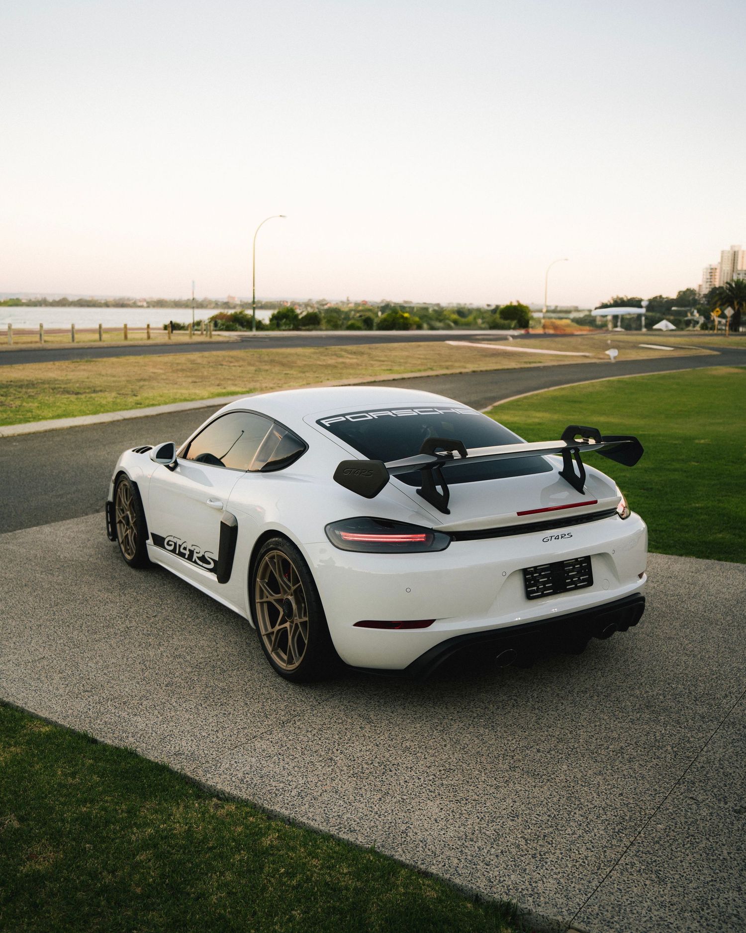 White Porsche GT sports car with large rear wing parked on waterfront road at sunset.