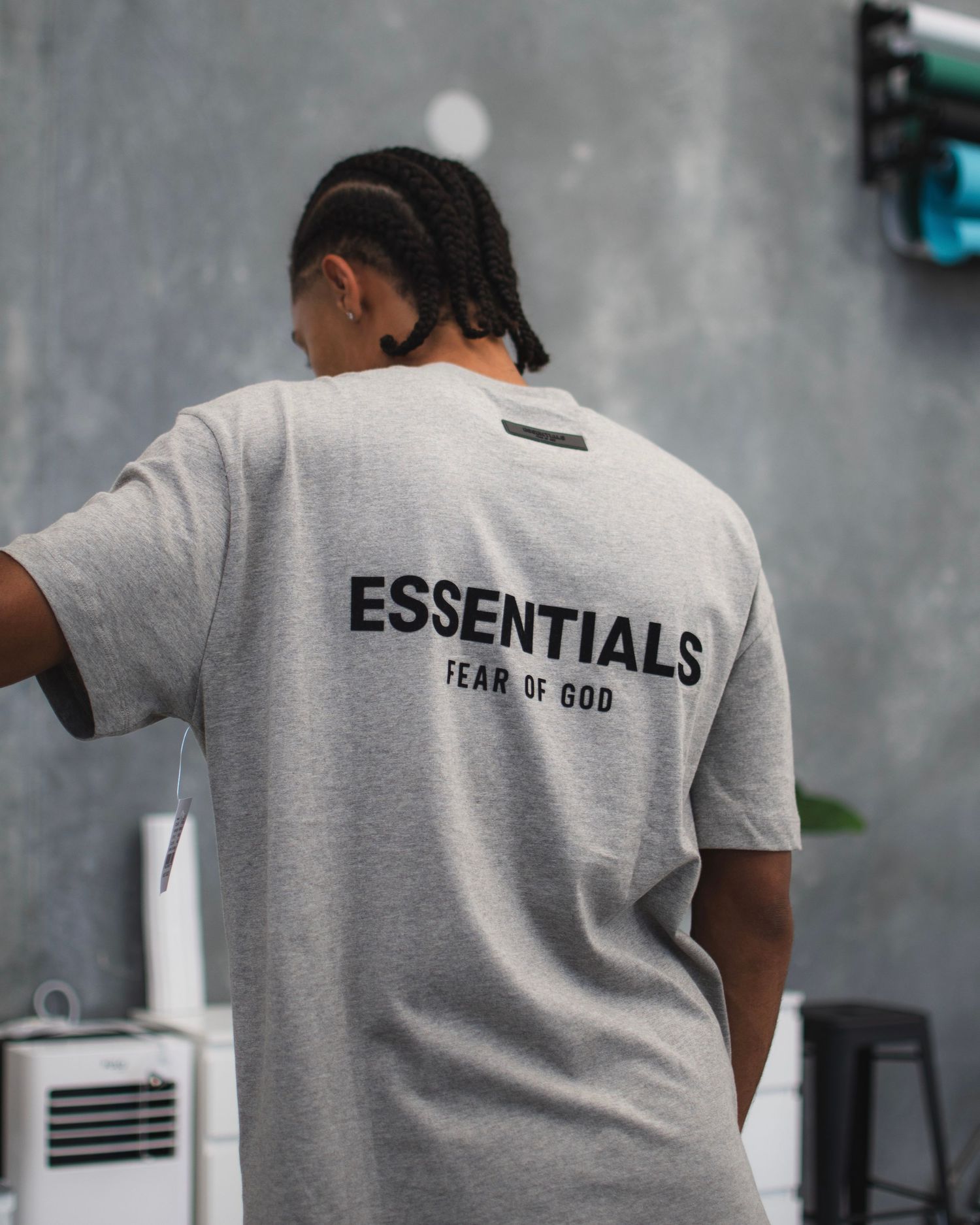Back view of grey t-shirt with 'ESSENTIALS FEAR OF GOD' text against concrete wall.