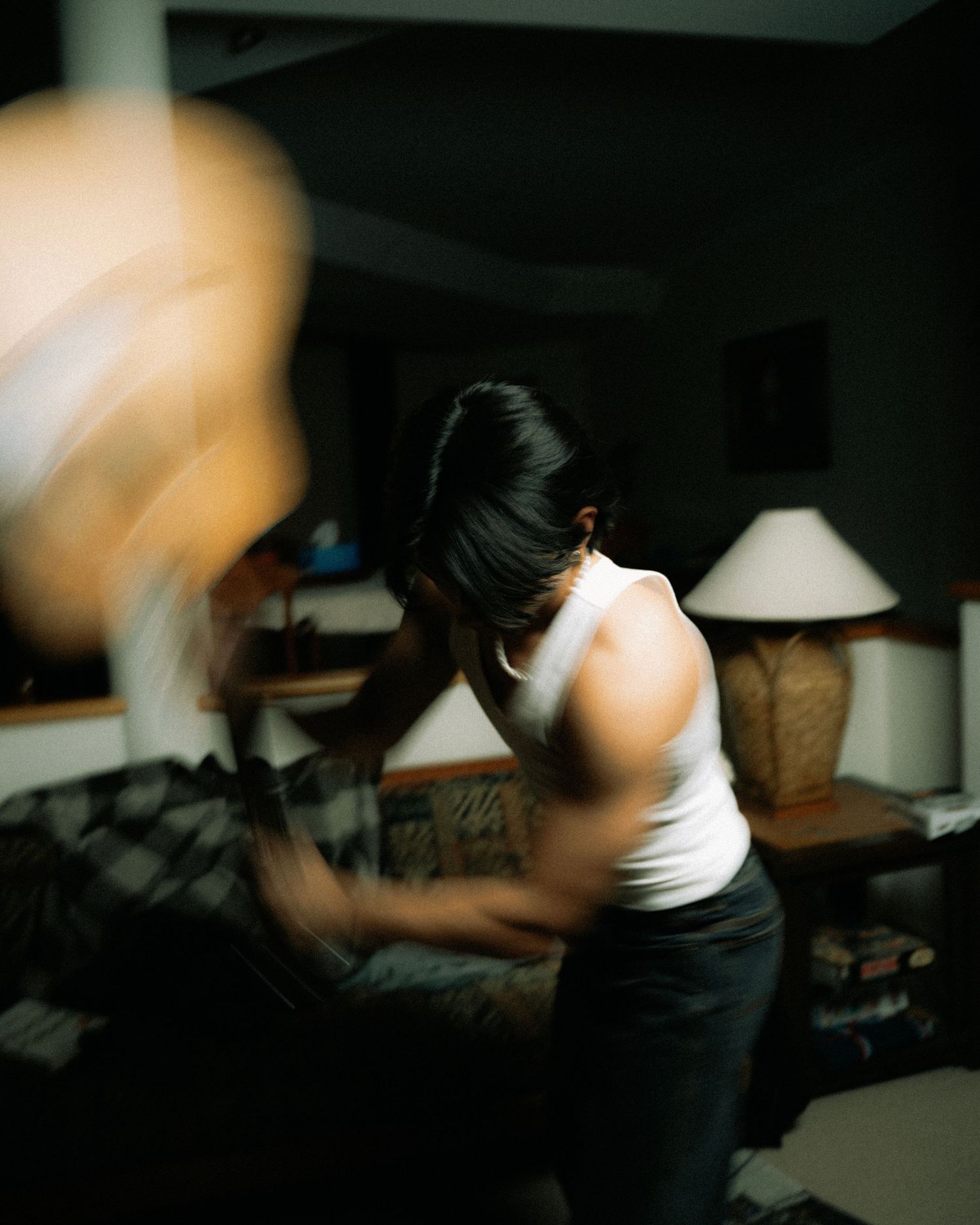 Motion-blurred figure in a white tank top exercises in a dimly lit room with table lamps visible in the background.