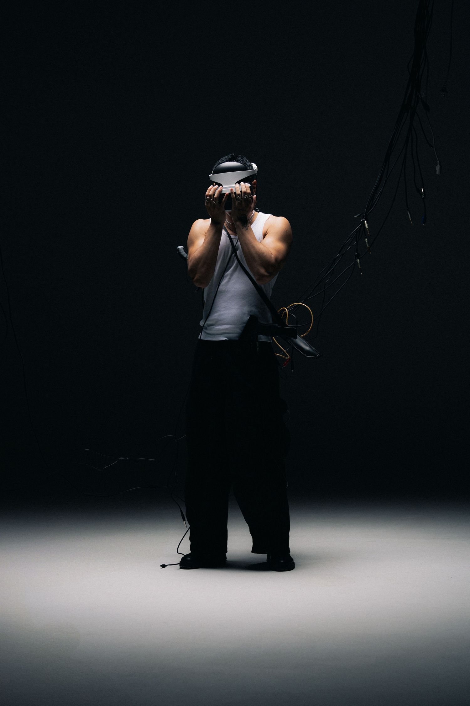 Photographer in gray tank top and dark pants holds camera to face in dramatic low-key lighting against dark background.