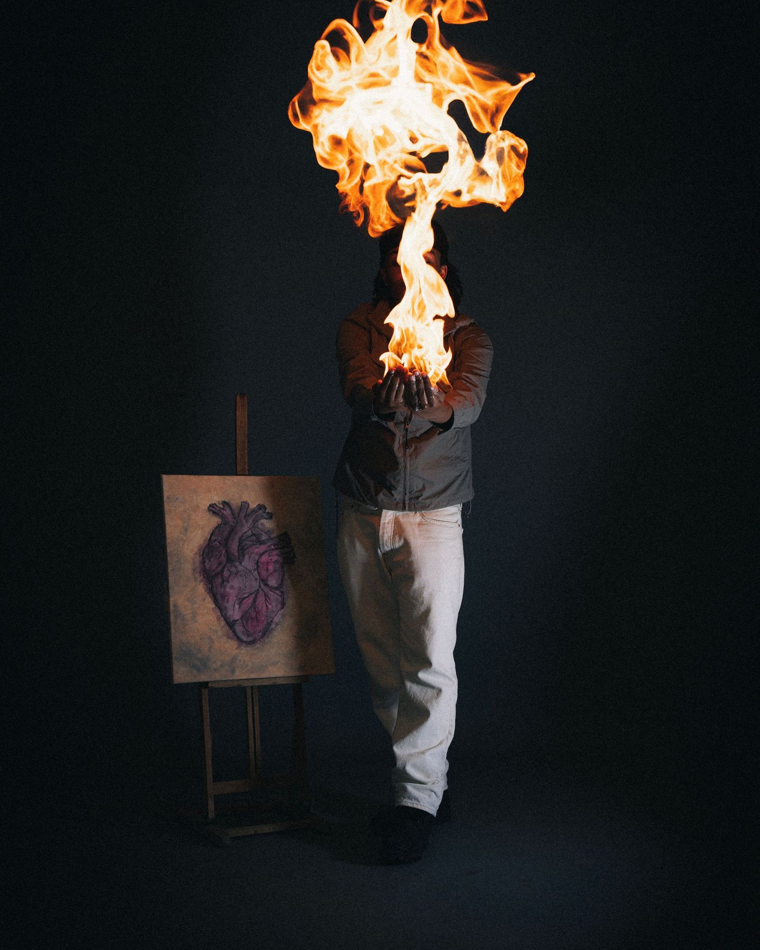 Artistic photograph of a figure with flames engulfing their head standing next to an easel with anatomical heart painting.