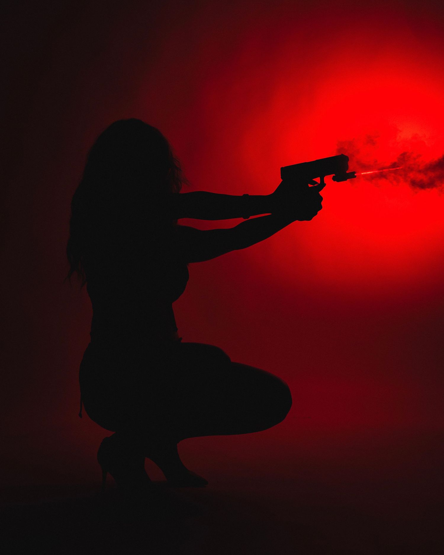 Silhouette of a person in crouching position firing a gun with dramatic red backlight creating smoke effects.