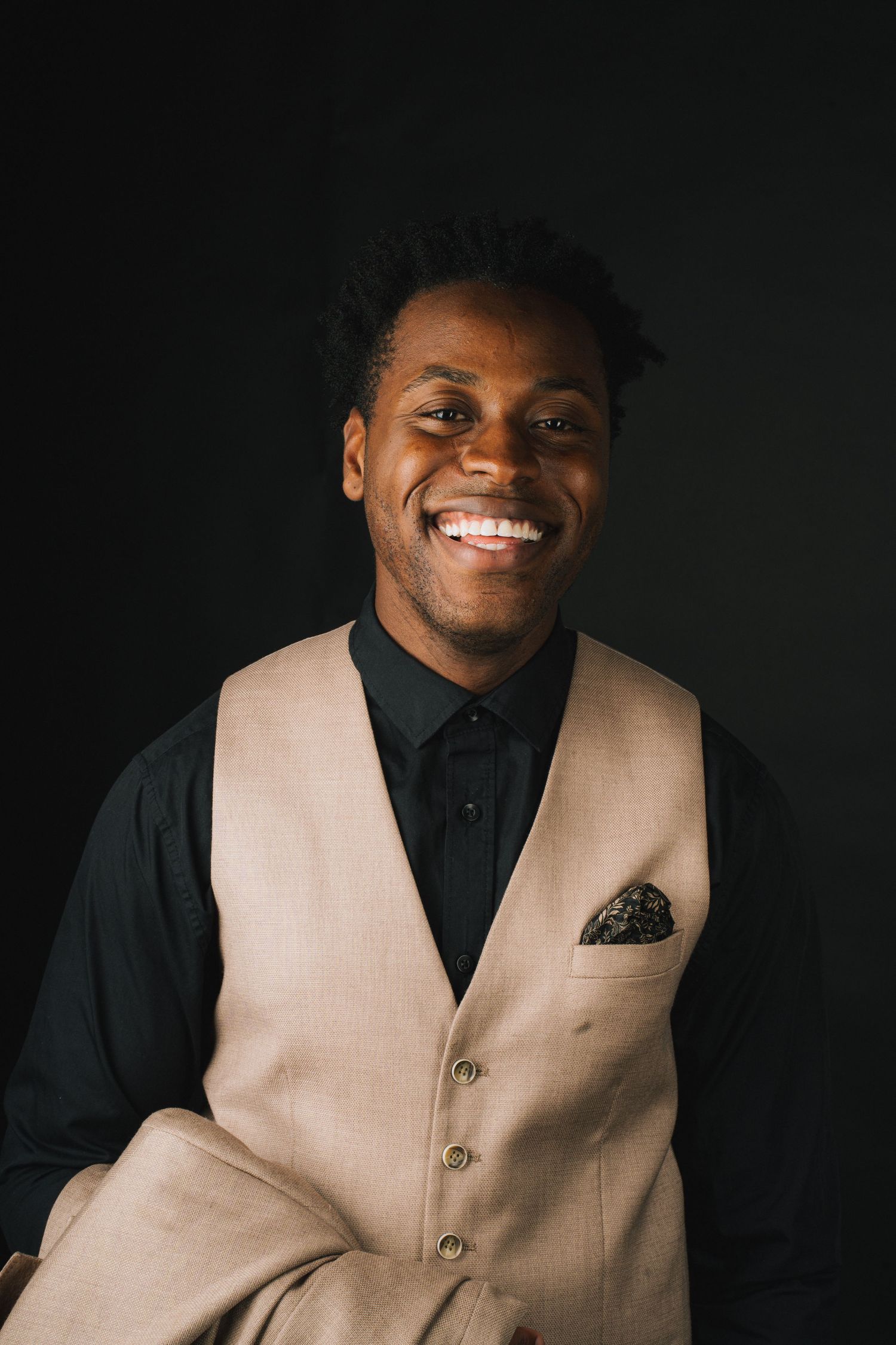 Professional portrait against dark background featuring warm smile, black dress shirt and tan vest with pocket square.