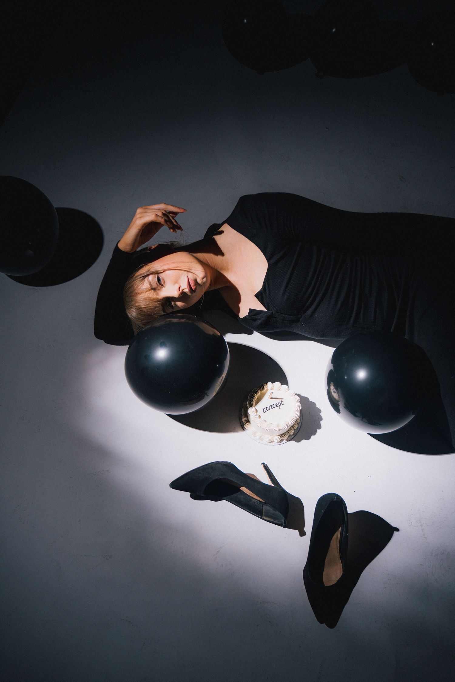 Artistic overhead shot showing a person in black lying on white floor with black balloons and shoes against dramatic lighting.