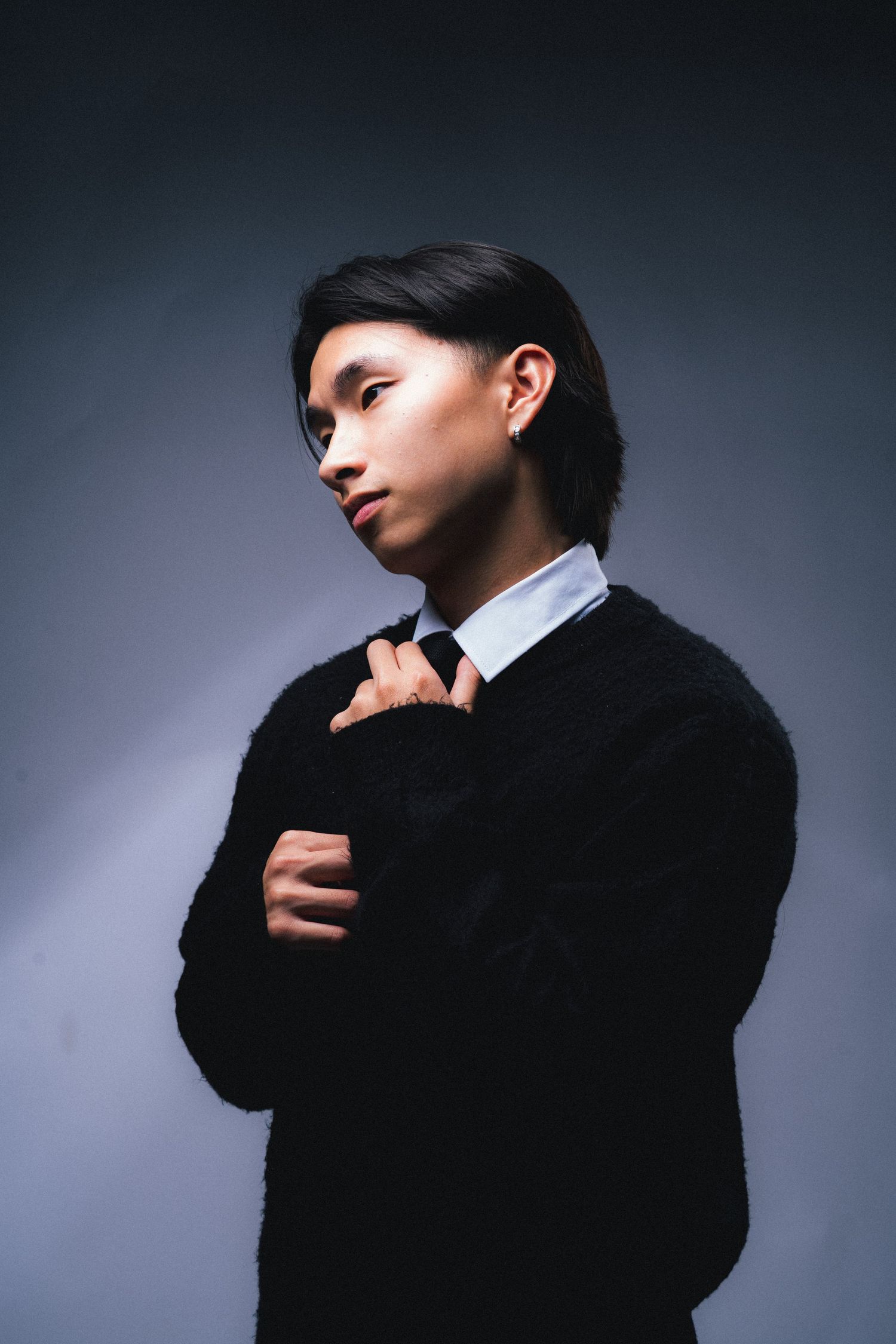 Dramatic portrait of a young Asian person in a black sweater adjusting their tie against a dark gradient background.