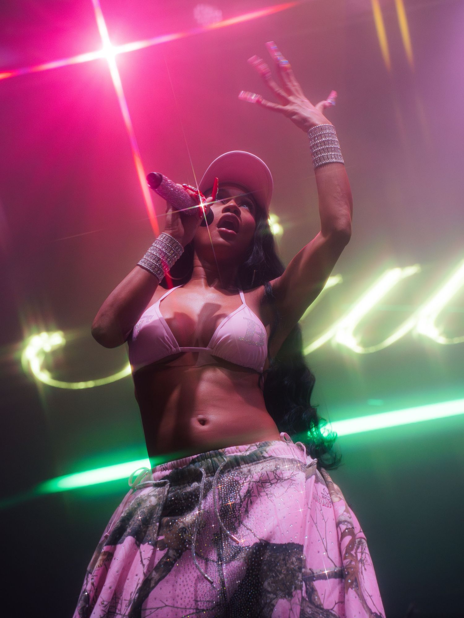 A performer in a white crop top and patterned skirt sings passionately on stage with pink and green lighting effects.