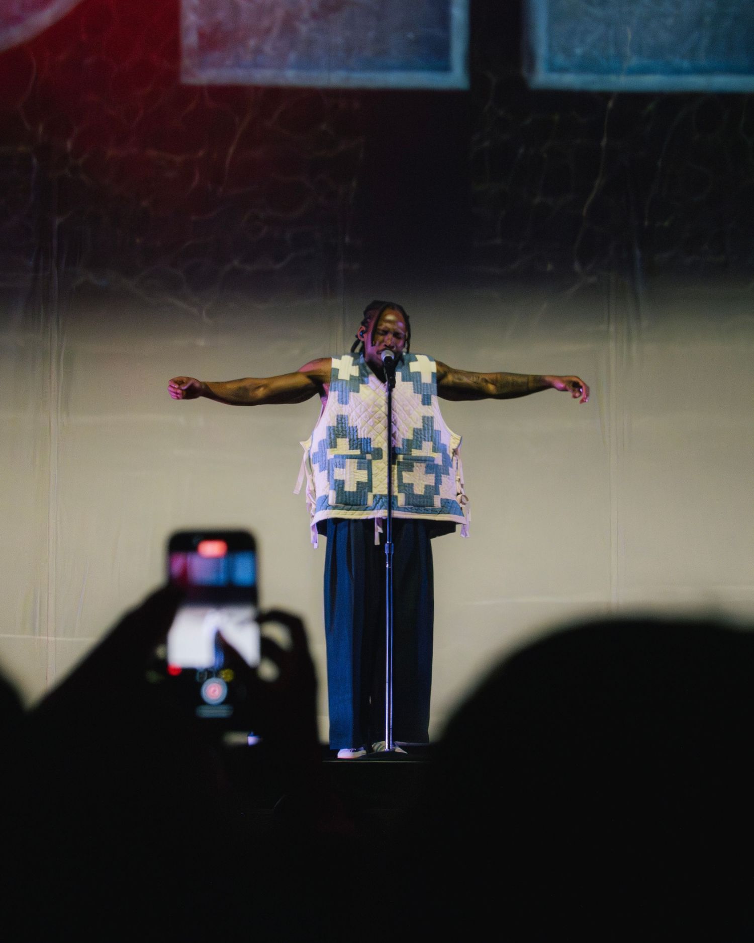 Performer in patterned vest strikes dramatic pose on stage with arms spread wide.