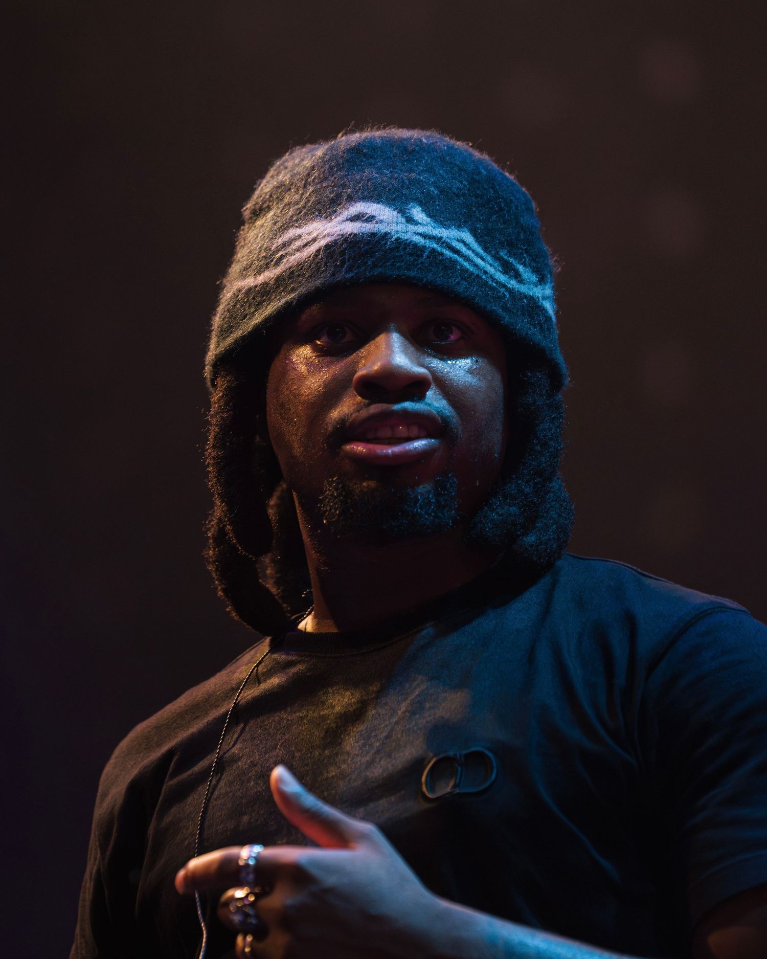 Performer in beanie and dark clothing photographed in dramatic low-key lighting on stage.