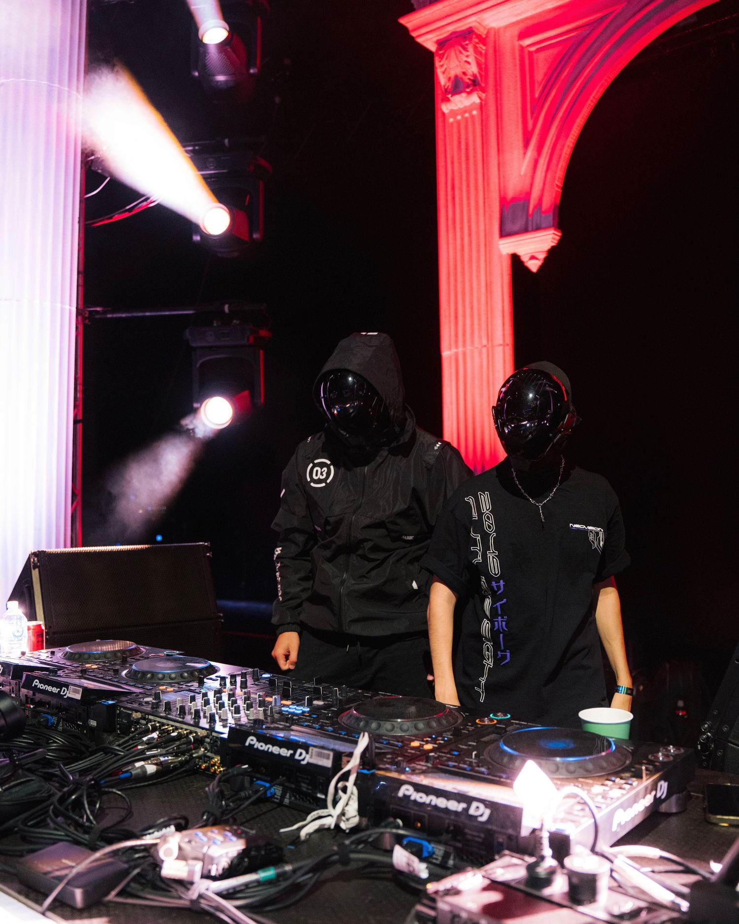 Two DJs in black hoodies perform at a venue with pink-lit architectural elements.