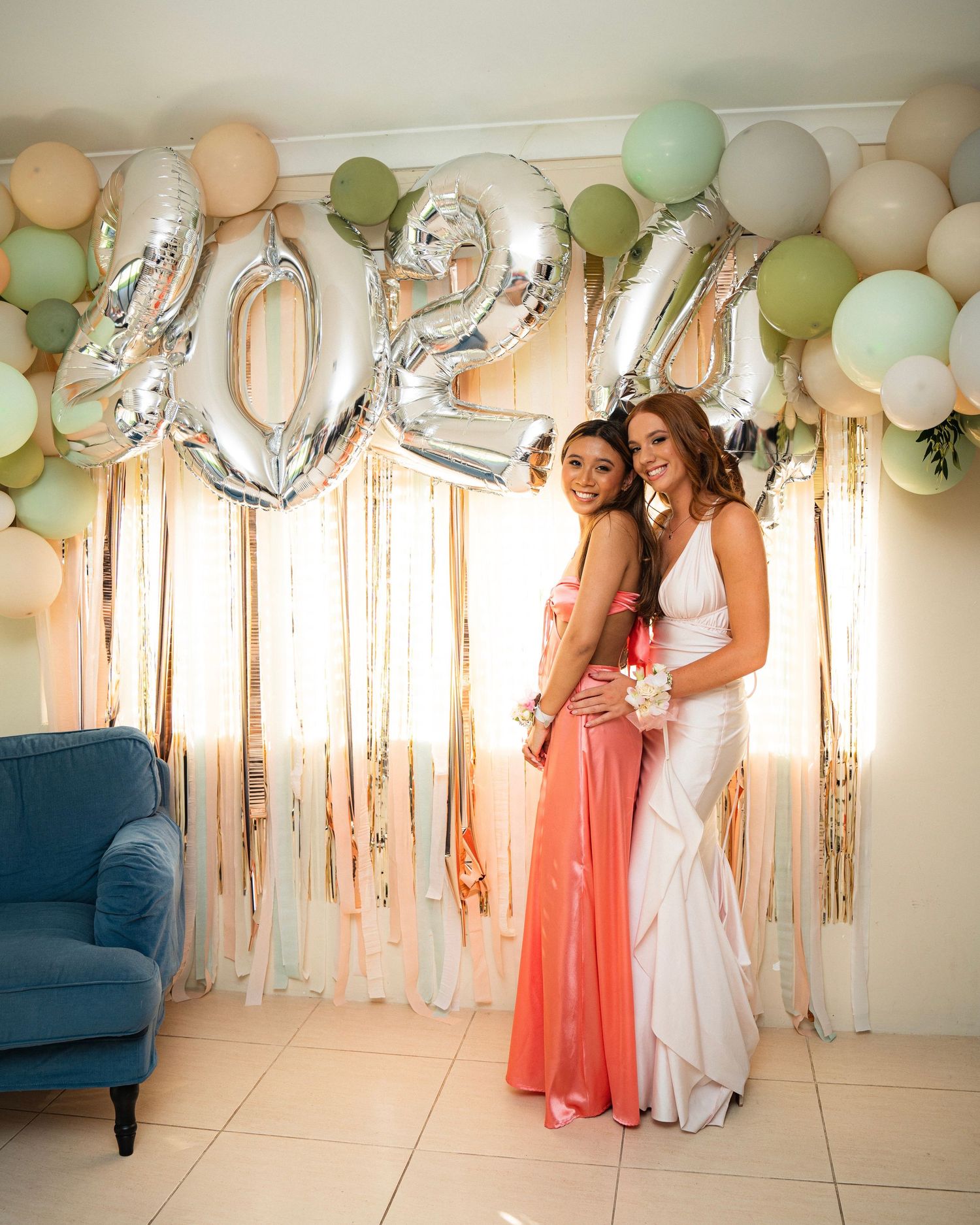 Two women in formal dresses posing under 2024 balloon decorations and pastel party balloons.