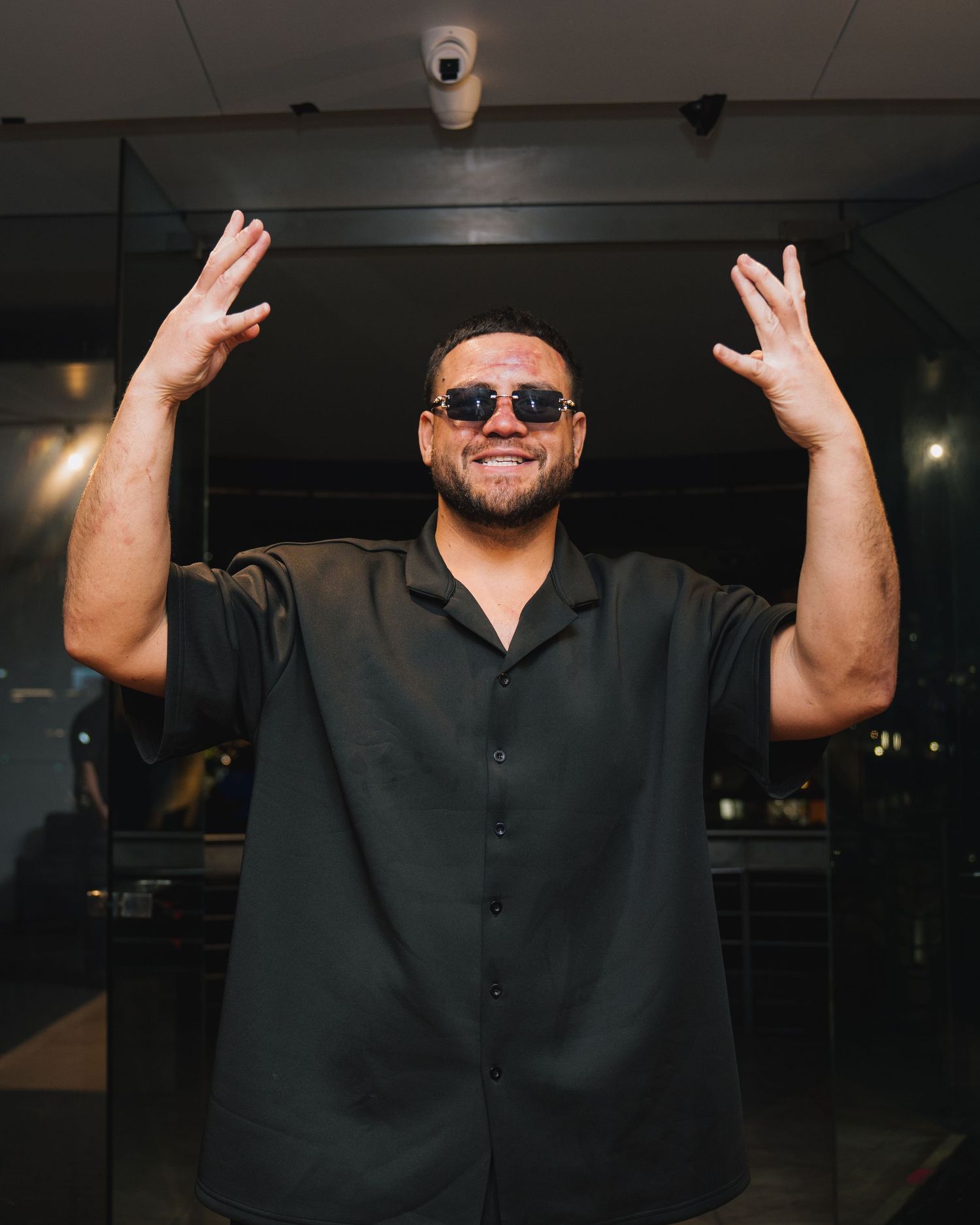 Man in black shirt raises both hands while smiling and wearing sunglasses indoors.