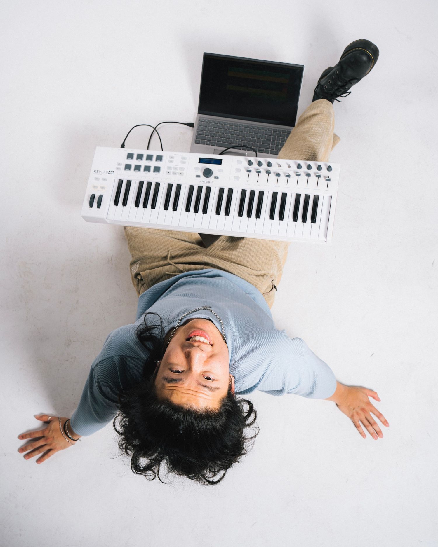 Overhead view of musician lying down with MIDI keyboard controller and laptop on white background.