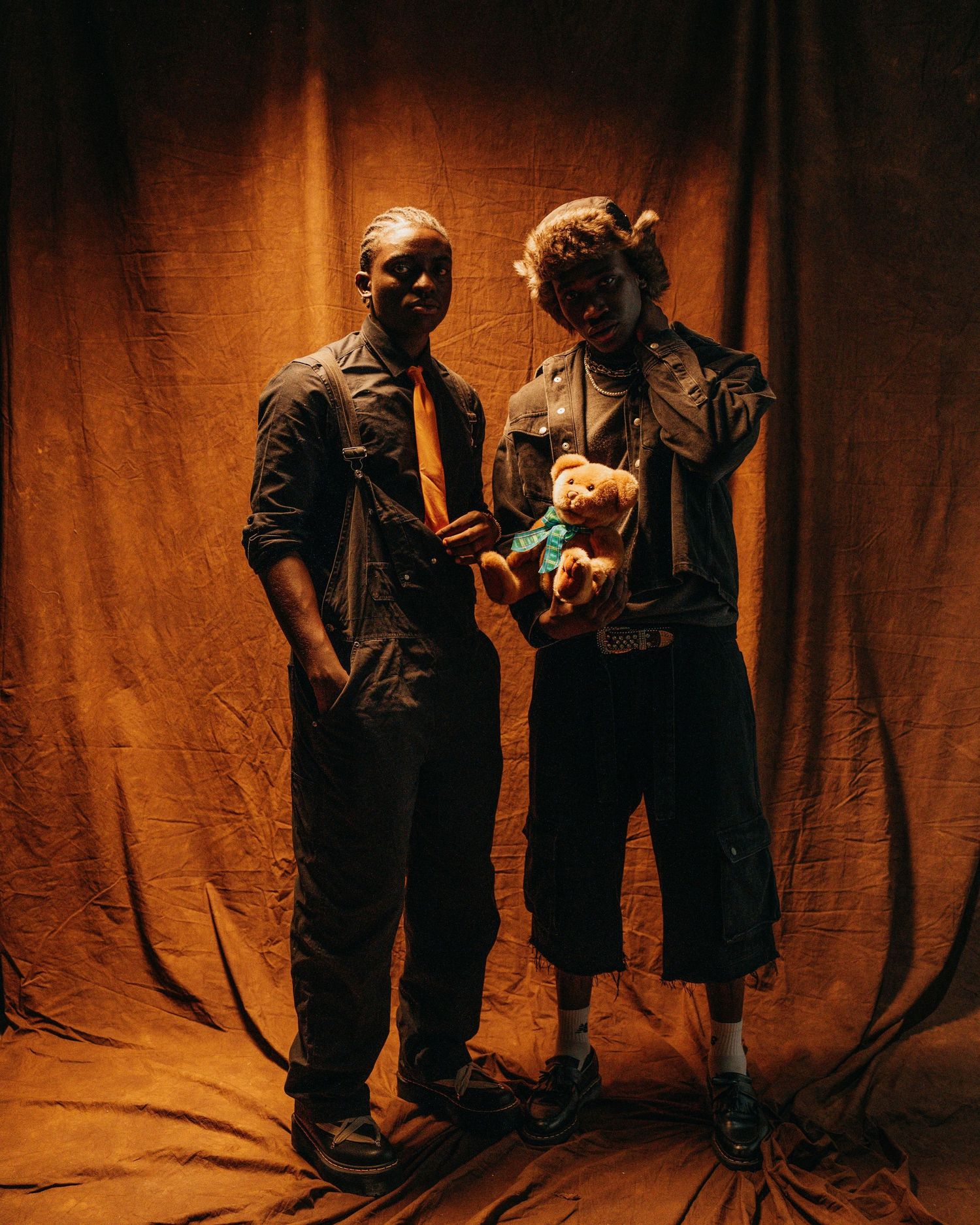 Two people in dark clothing pose with stuffed toy against orange-lit backdrop.