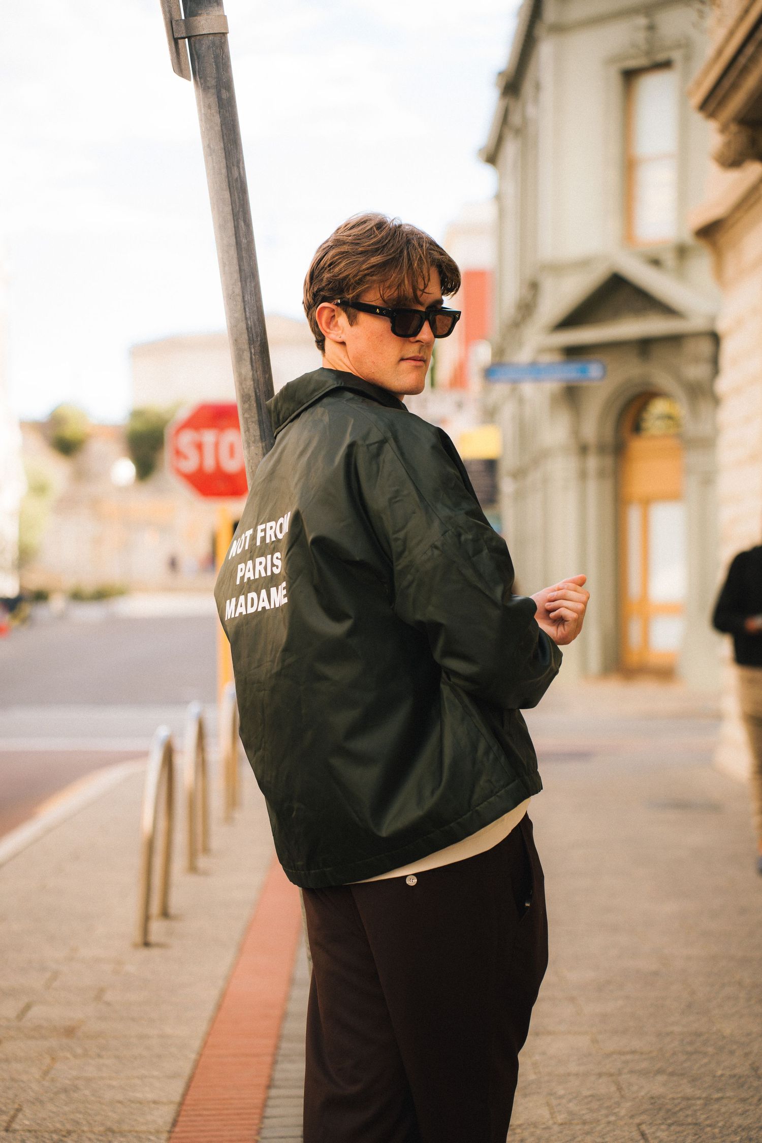 Person in dark jacket and sunglasses standing on city sidewalk next to stop sign.