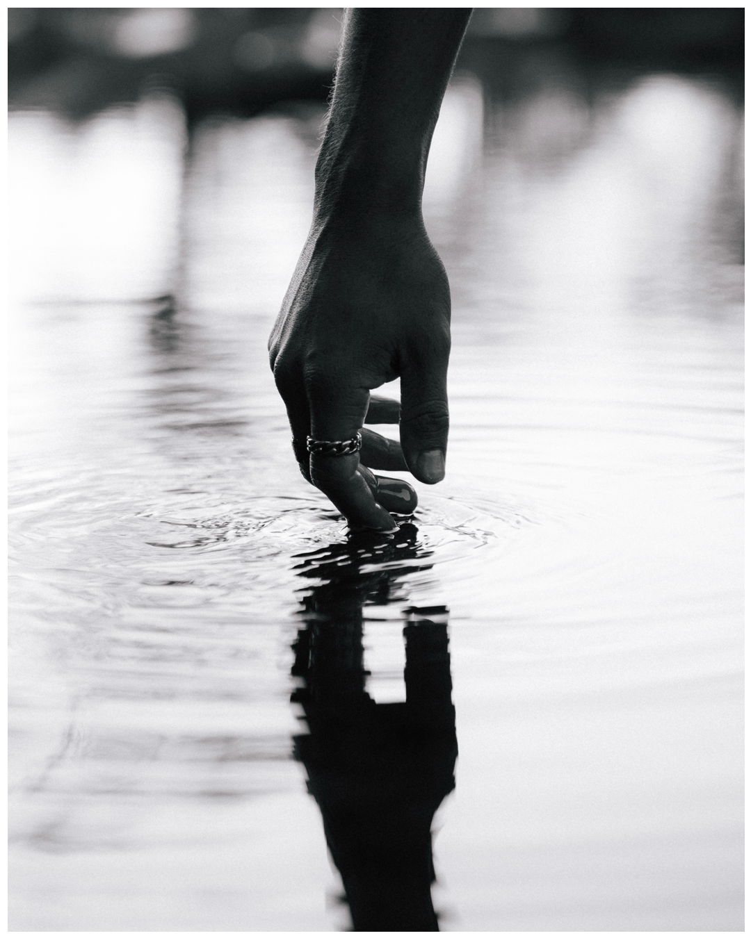 A hand gently touches the surface of still water creating a reflection in dramatic black and white photography.