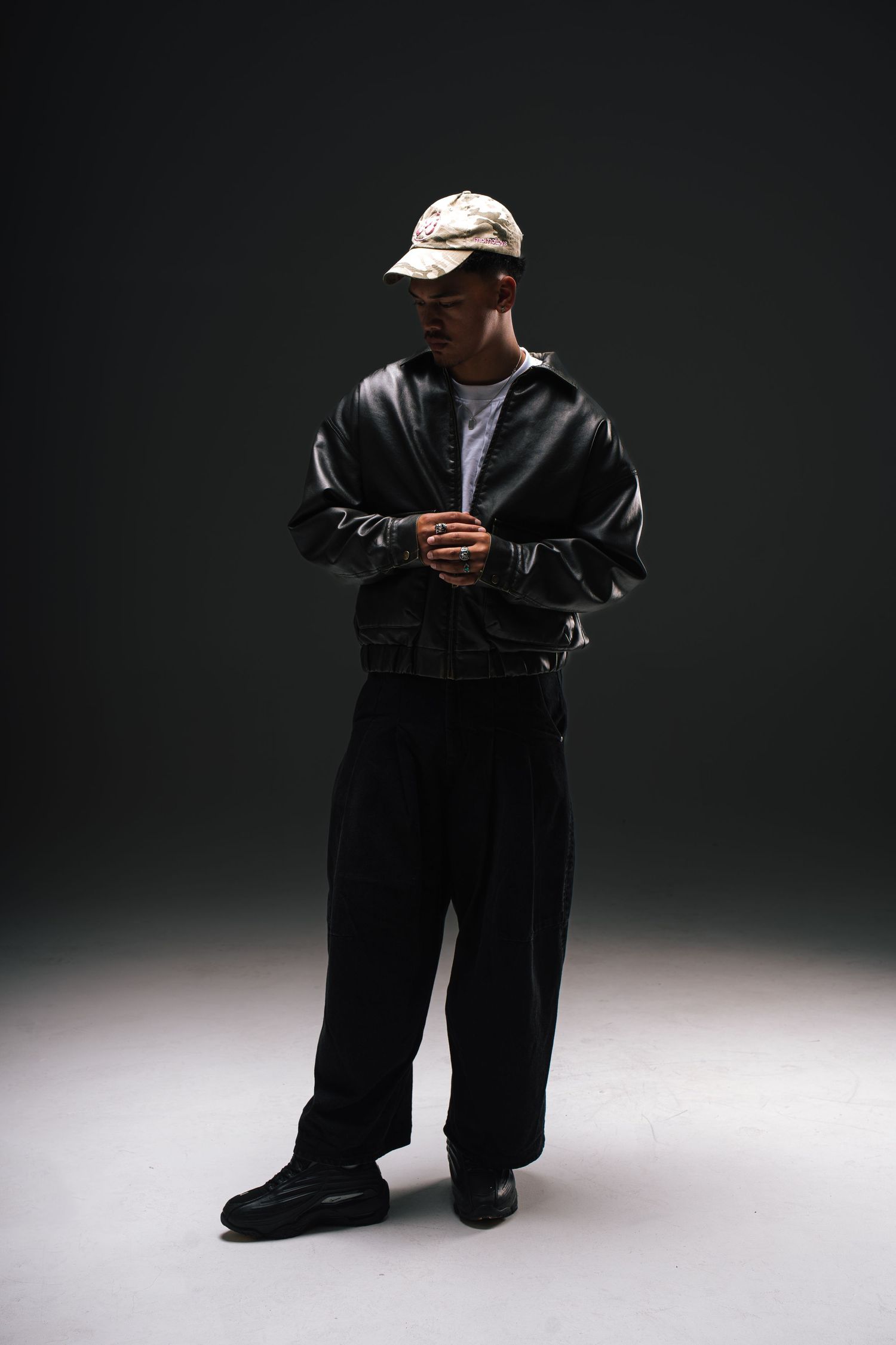 Person wearing dark casual outfit with white cap and jacket posing against dark background in dramatic lighting.