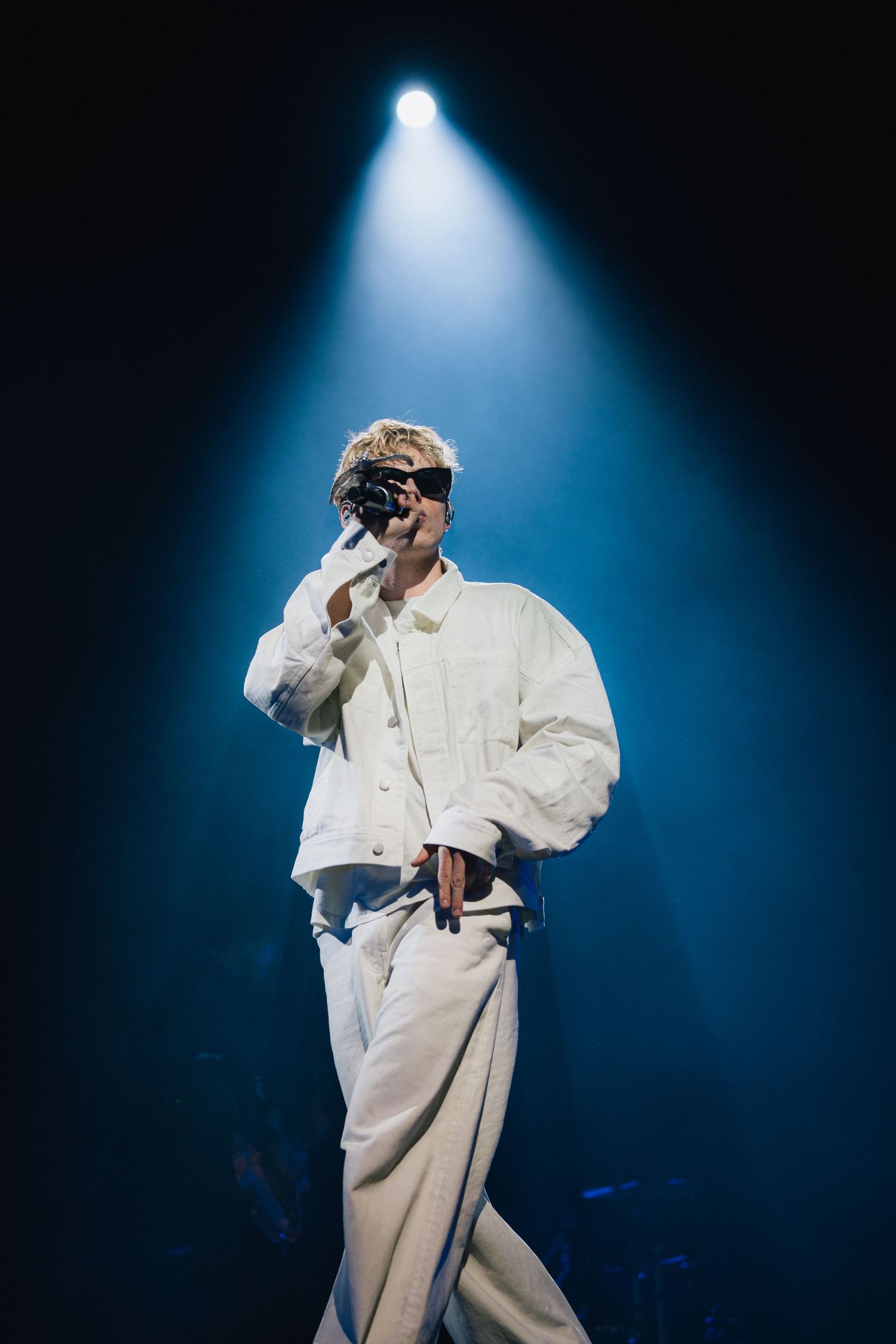 Performer in all-white outfit singing on stage under dramatic blue spotlight during concert performance.