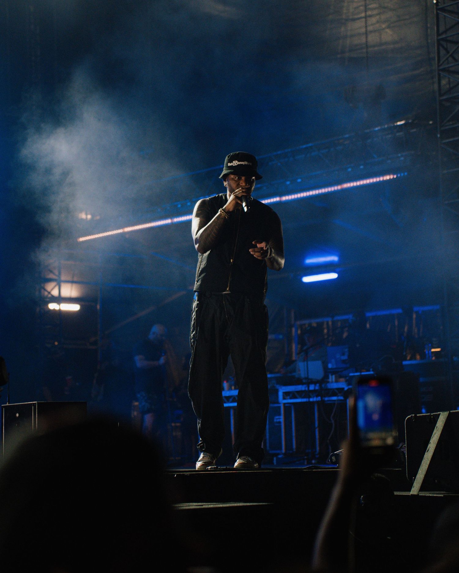 Performer in dark clothing singing on smoky stage with blue lighting effects.
