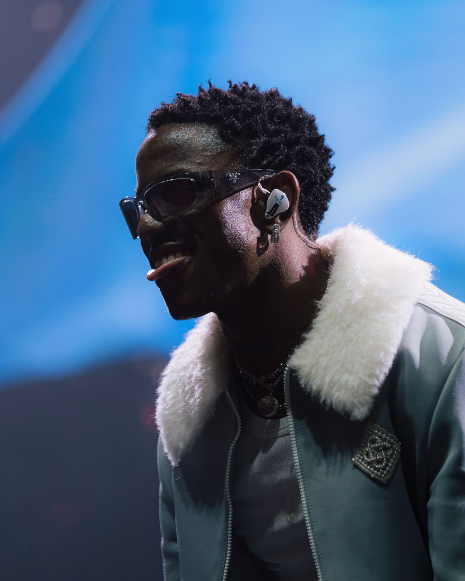 Performer wearing sunglasses and white fur collar jacket smiling during outdoor concert performance.