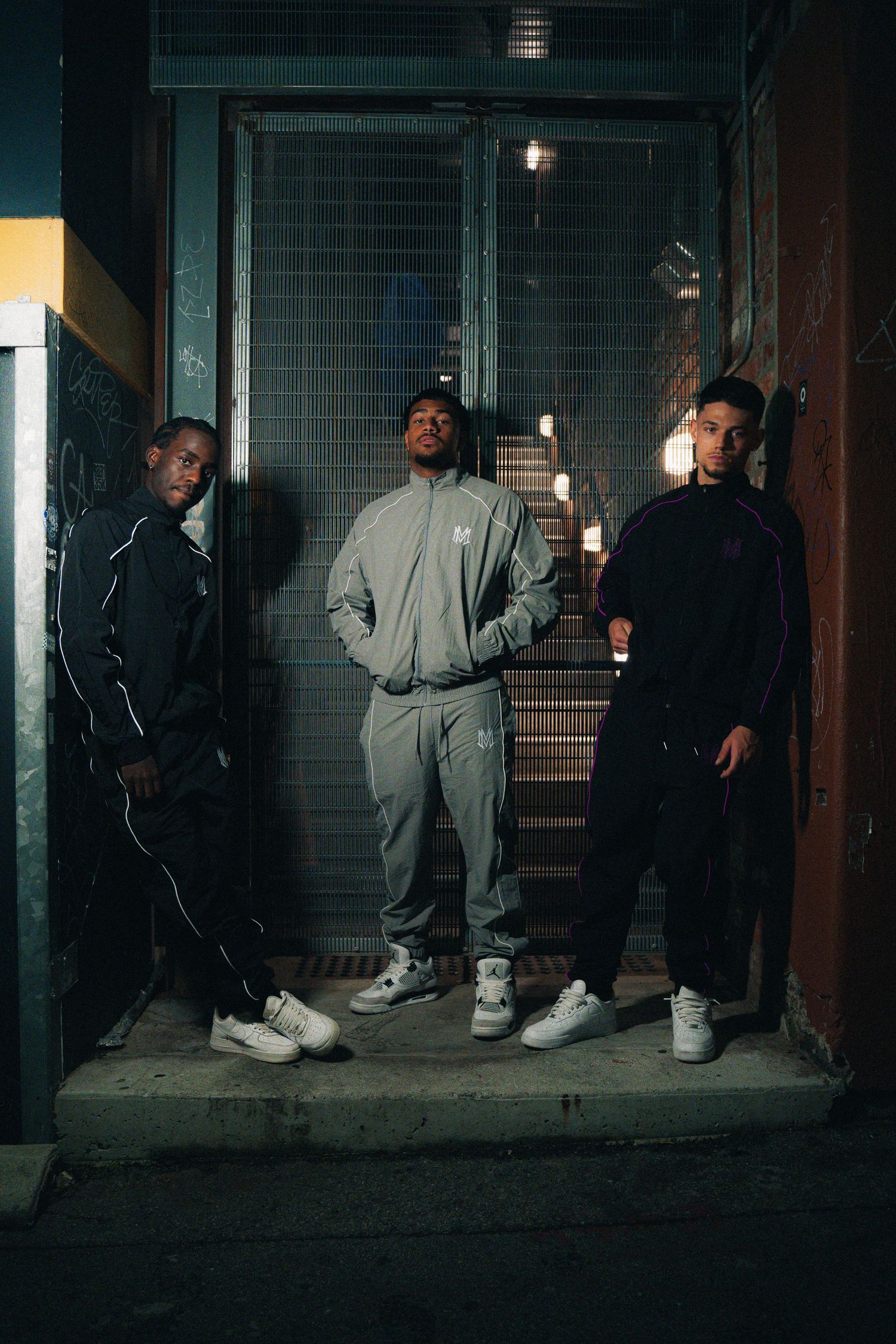 Three people in streetwear stand on dimly lit steps in urban setting.