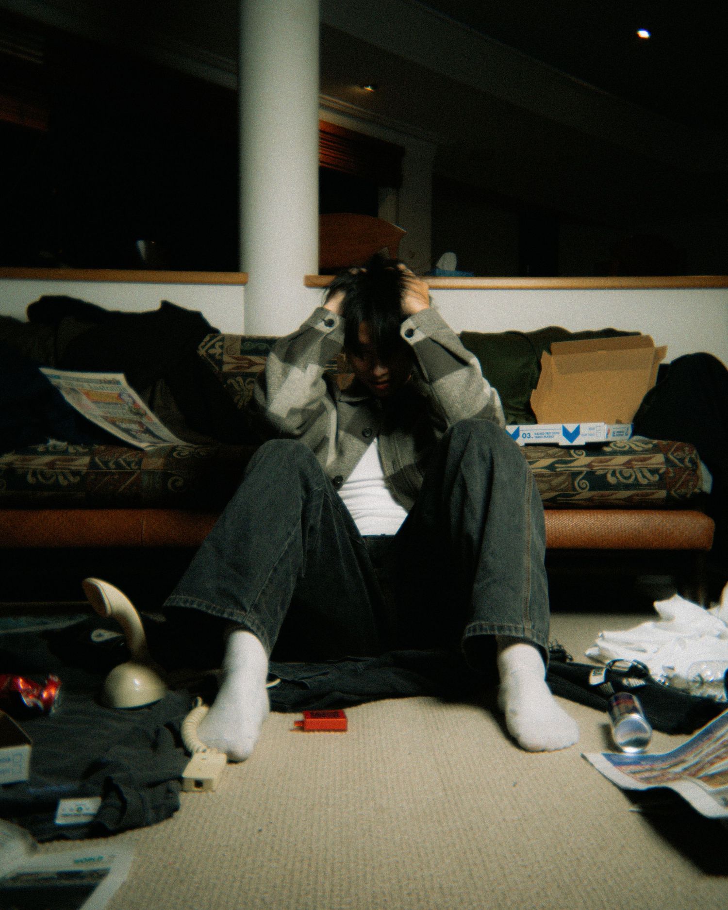 Person in casual clothing sitting on carpet surrounded by scattered clothes and papers in dimly lit room.