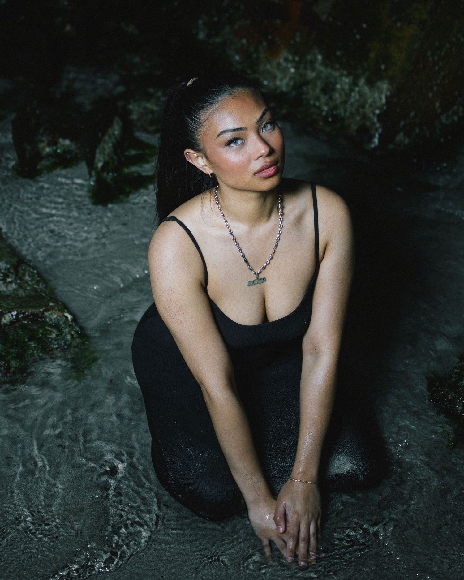 A person wearing a black dress and necklace poses thoughtfully in a dark natural setting with moody lighting.