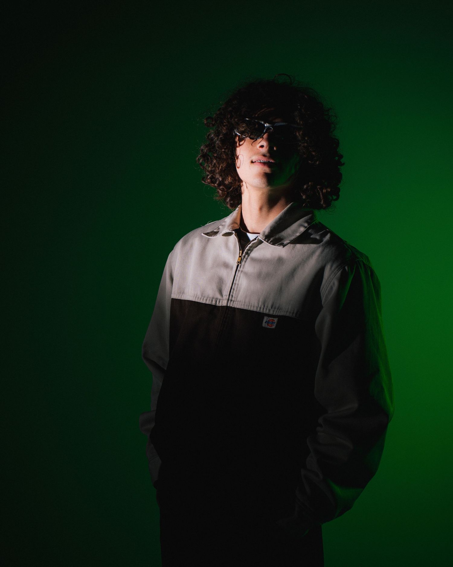 Portrait of a person with curly black hair wearing a two-tone jacket against a dark green backdrop.