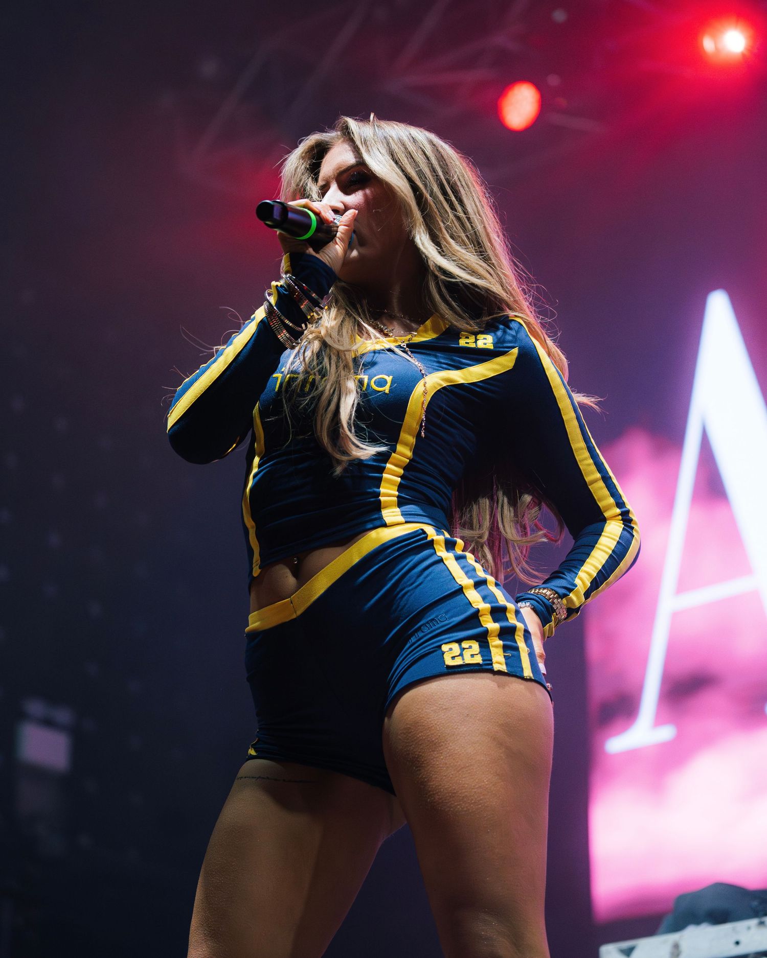 A performer in a blue and yellow athletic outfit sings into a microphone on stage with red lighting effects.