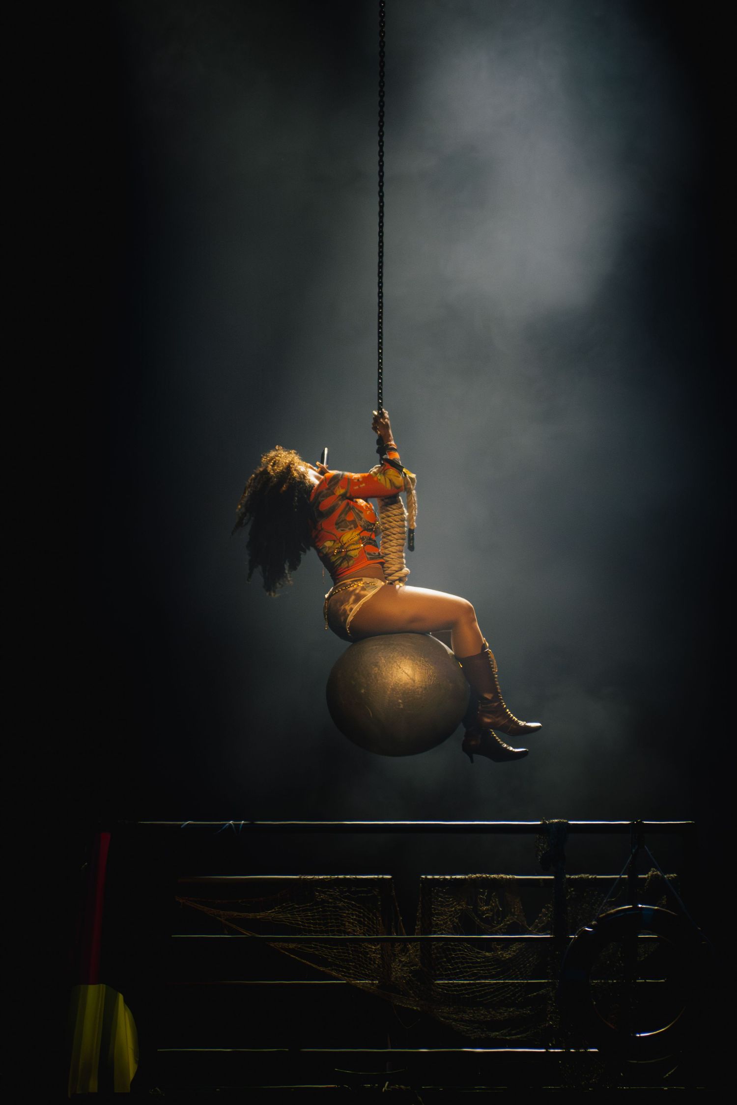 A circus performer in an orange costume swings through dramatic lighting while balancing on a large metallic sphere.