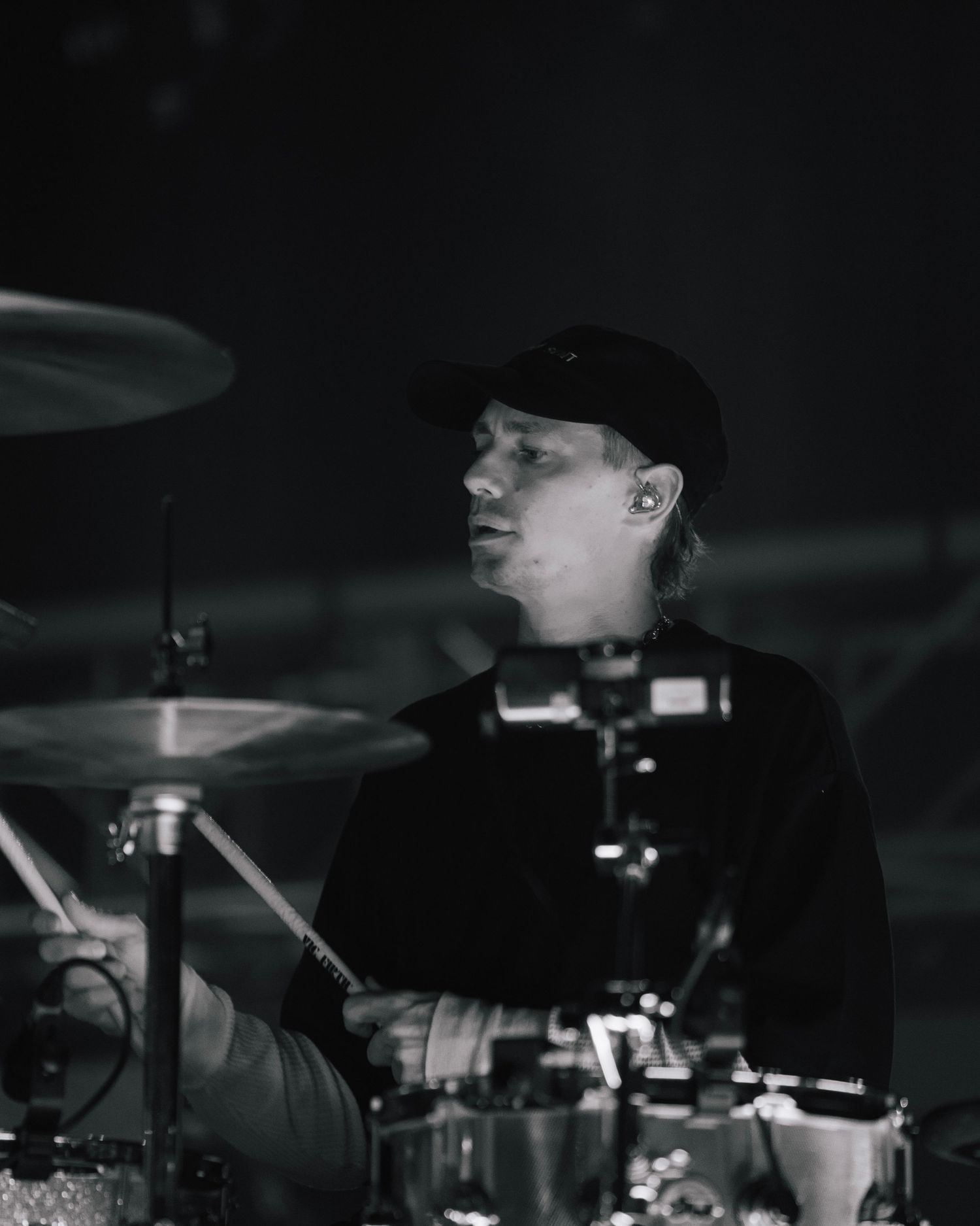 Black and white photograph of a drummer performing behind a drum kit in low lighting.