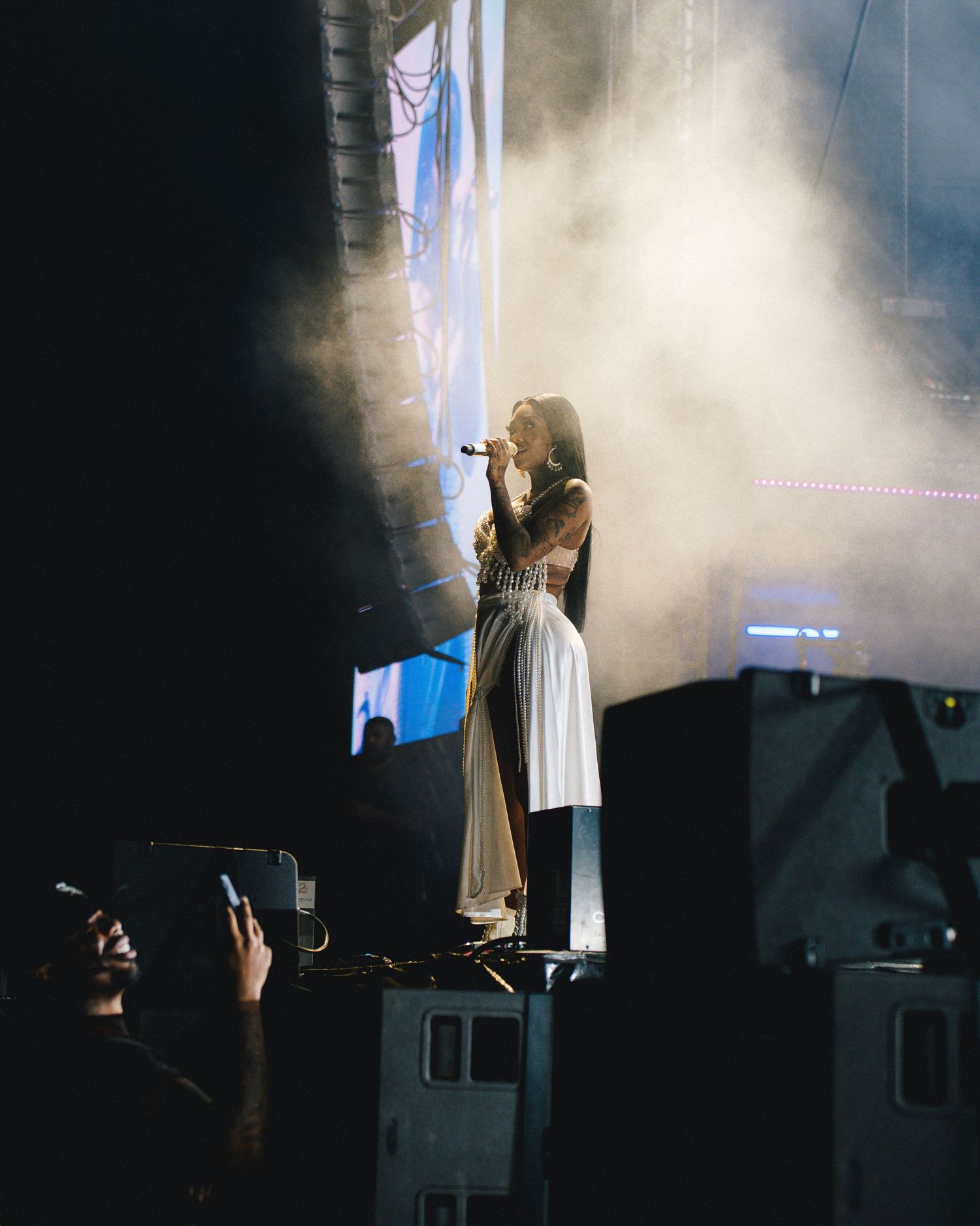 A performer in a white dress stands on stage in dramatic spotlight and fog during a concert performance.