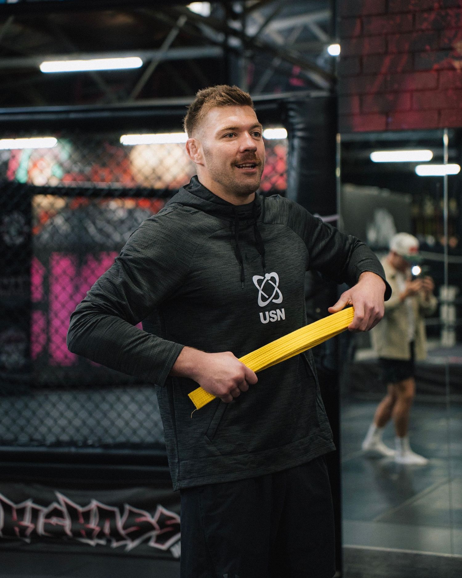 Fitness instructor holding yellow equipment in modern gym setting with cage background.