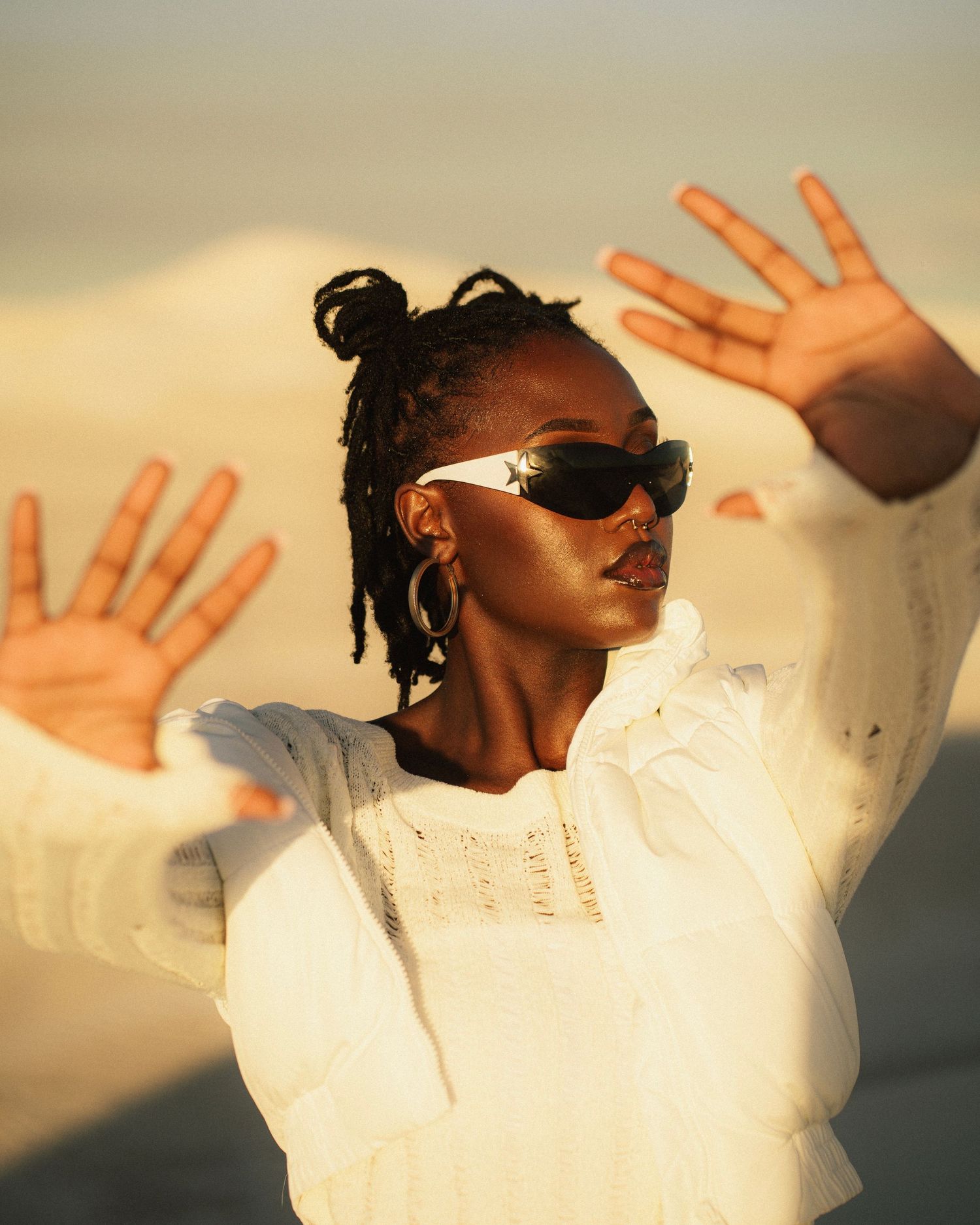 Person in white sweater raising hands while wearing sunglasses in golden desert light.