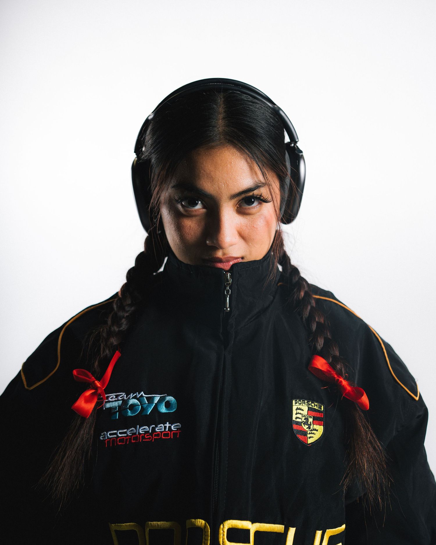 Person in black Porsche jacket wearing headphones and braided hair against white background.