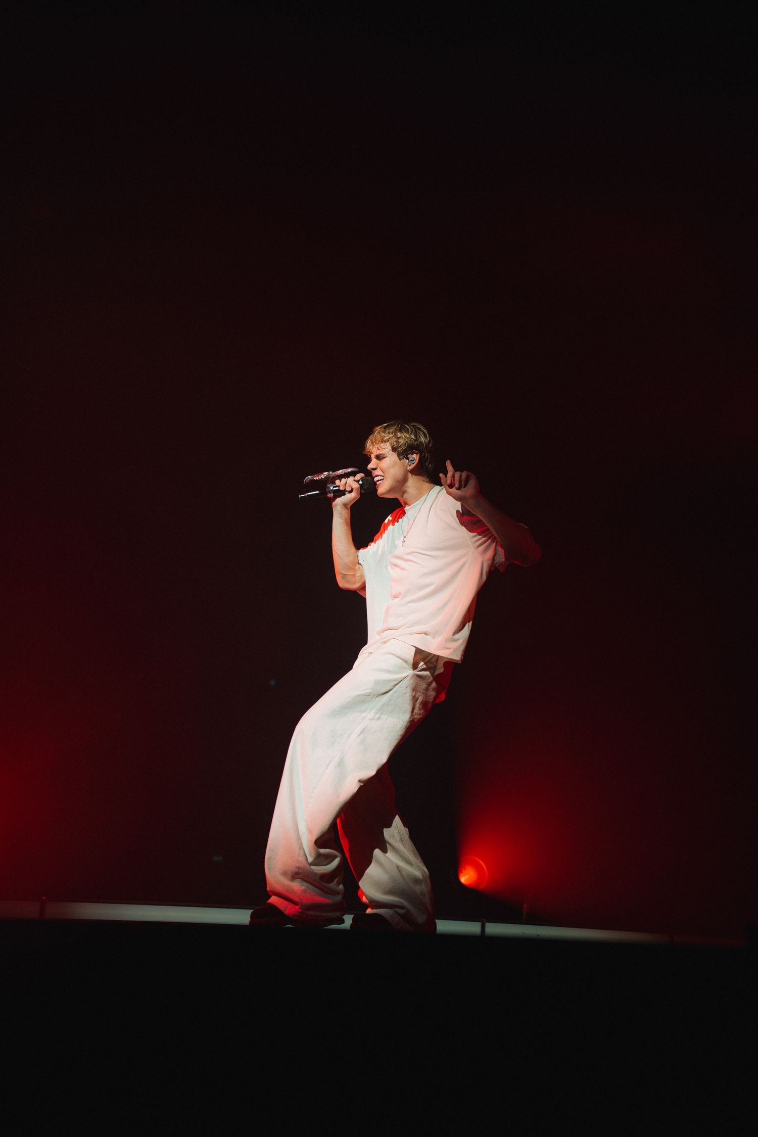 A performer in all white attire energetically performs on stage with a microphone against dark red backdrop.