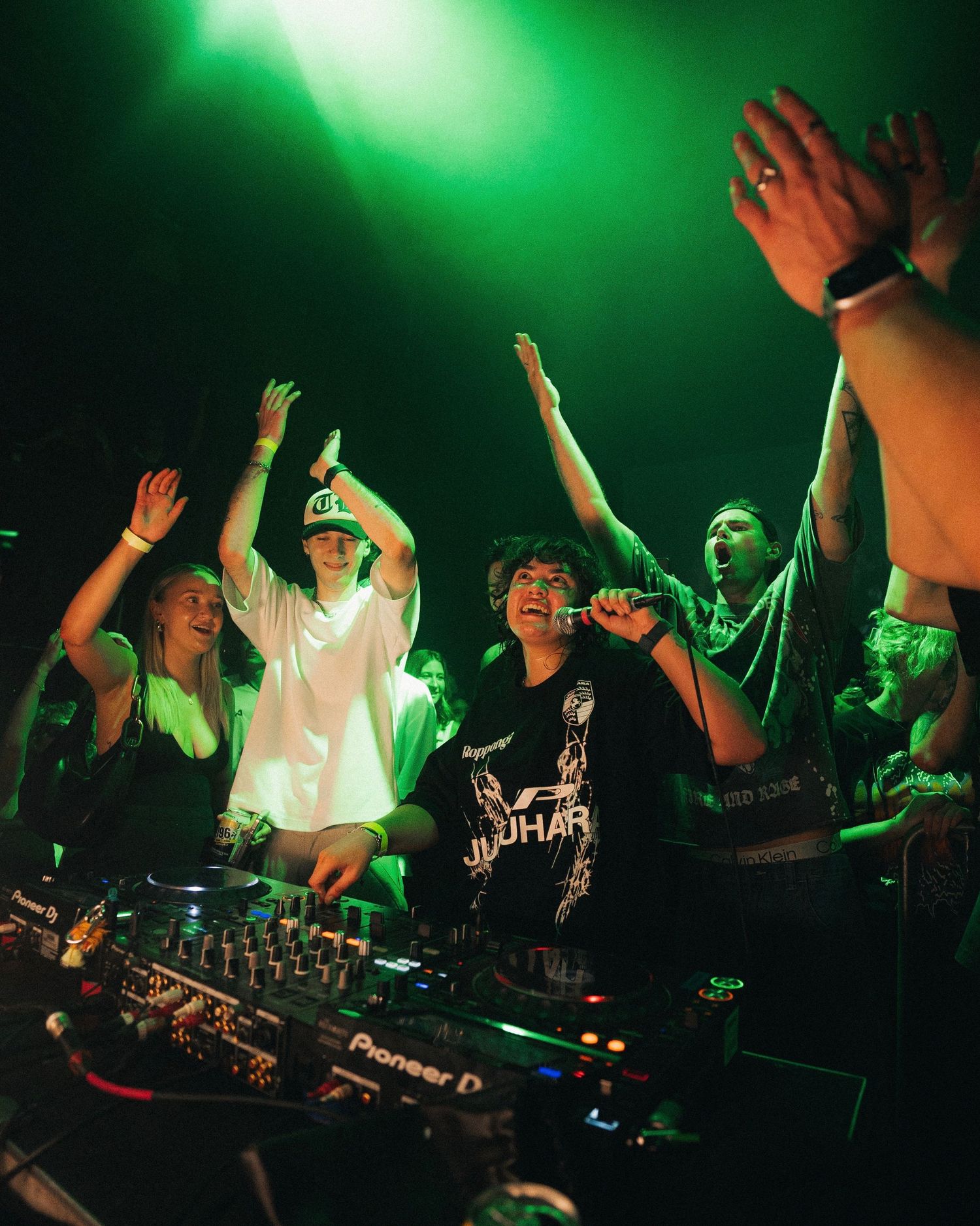 Energetic crowd with raised hands at concert venue under green stage lighting.